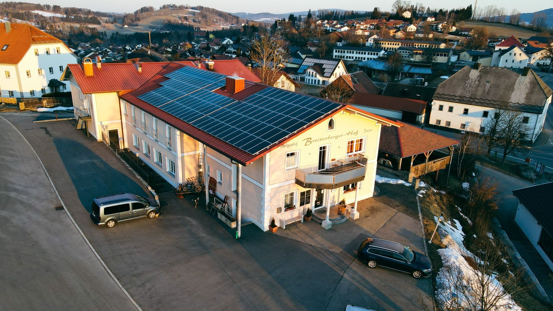 Luftaufnahme des Breitenberger Hofs*** mit Sonnenkollektoren und einem Wellnessbereich auf dem Dach, umgeben von geparkten Autos und Häusern in einer kleinen, hügeligen Stadt bei Passau. Auf dem Boden sind Schneeflecken zu sehen.