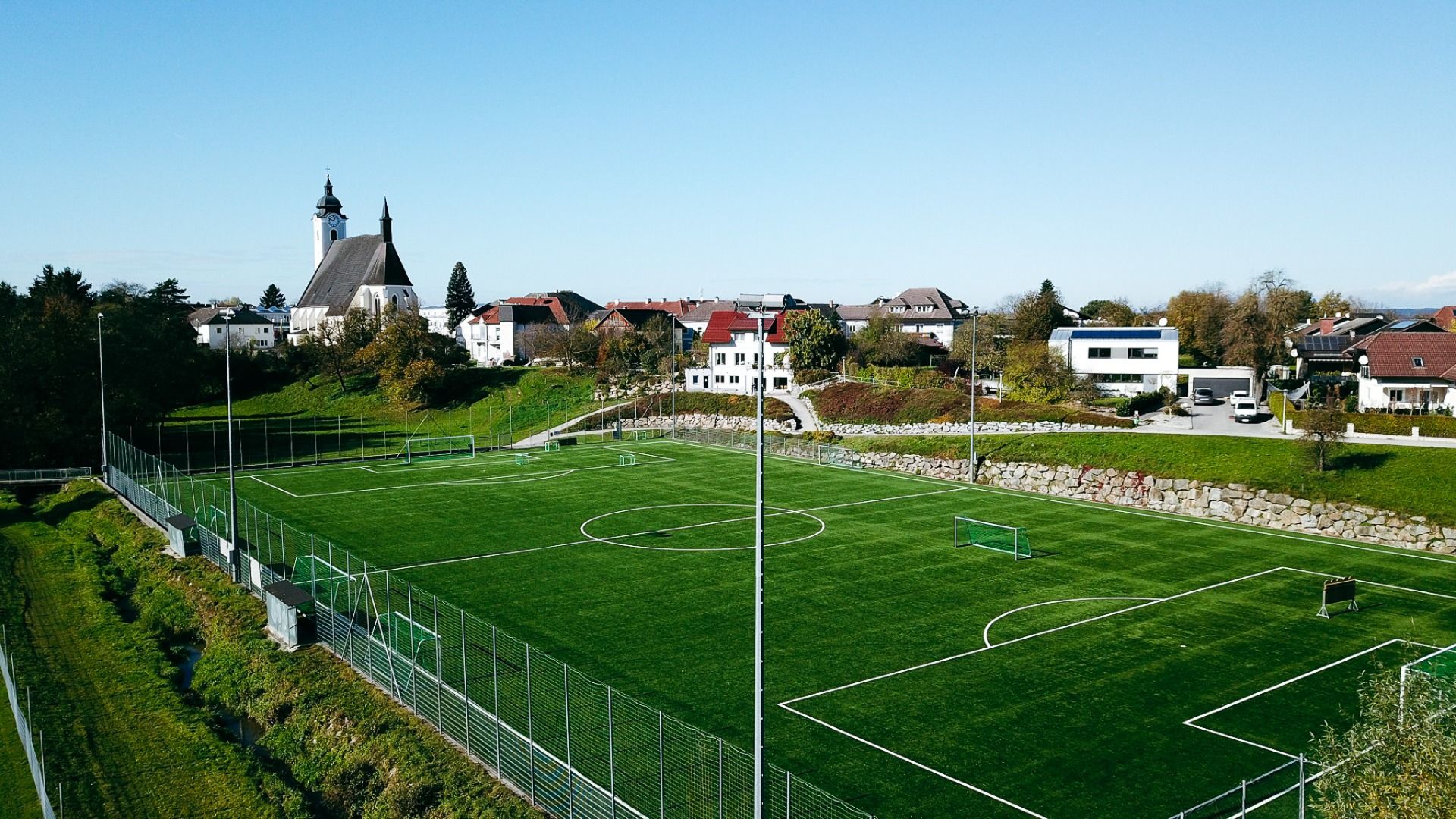 Flyfoto av den grønne kunstgressbanen i Neuhofen an der Ybbs, i nærheten av hotellet Perbersdorfer Heurigen. Omgitt av et gjerde med små porter, hus og et stort kirketårn under en knallblå himmel.