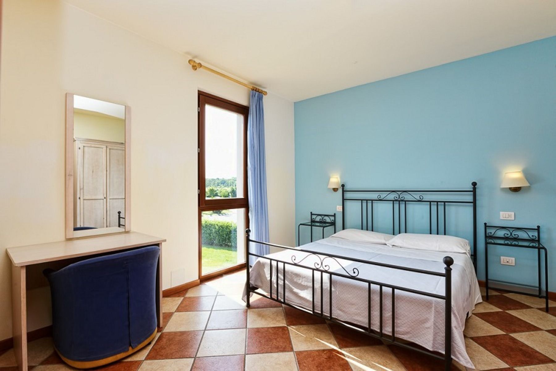 Helles Schlafzimmer in der Appartements Residence Eden*** mit schwarzem Metallbettgestell, weißer Bettwäsche, blauer Akzentwand, gefliestem Boden, Holzschreibtisch mit Spiegel, blauem Stuhl und Fenster mit blauem Vorhang. Ideal für preisbewusste Mannschaf