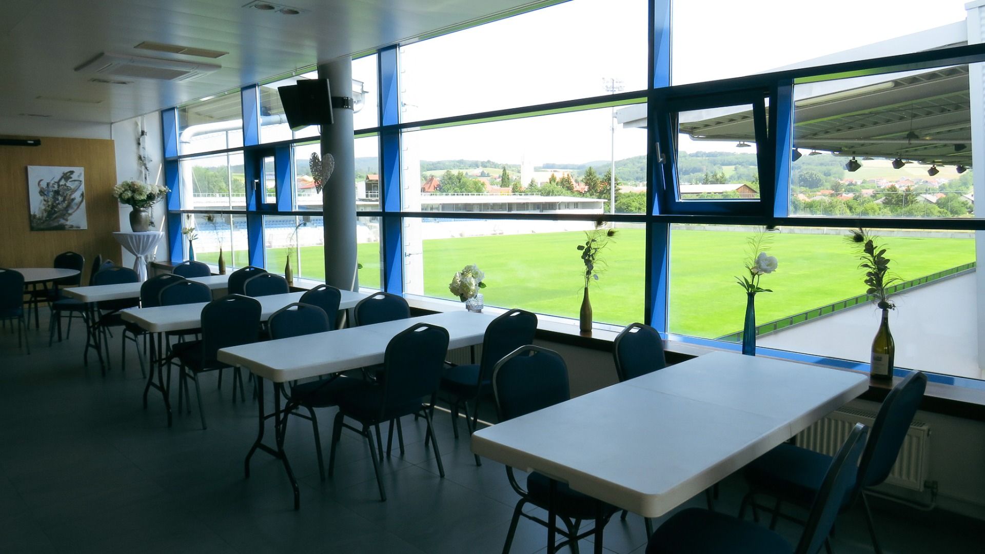 Der moderne Innenbereich im Gasthof & Gästehaus Trummer ist mit blauen Stühlen, weißen Tischen mit Blumenvasen und großen Glasfenstern mit Blick auf den Kunstrasenplatz der Sportanlage Ritzing ausgestattet.