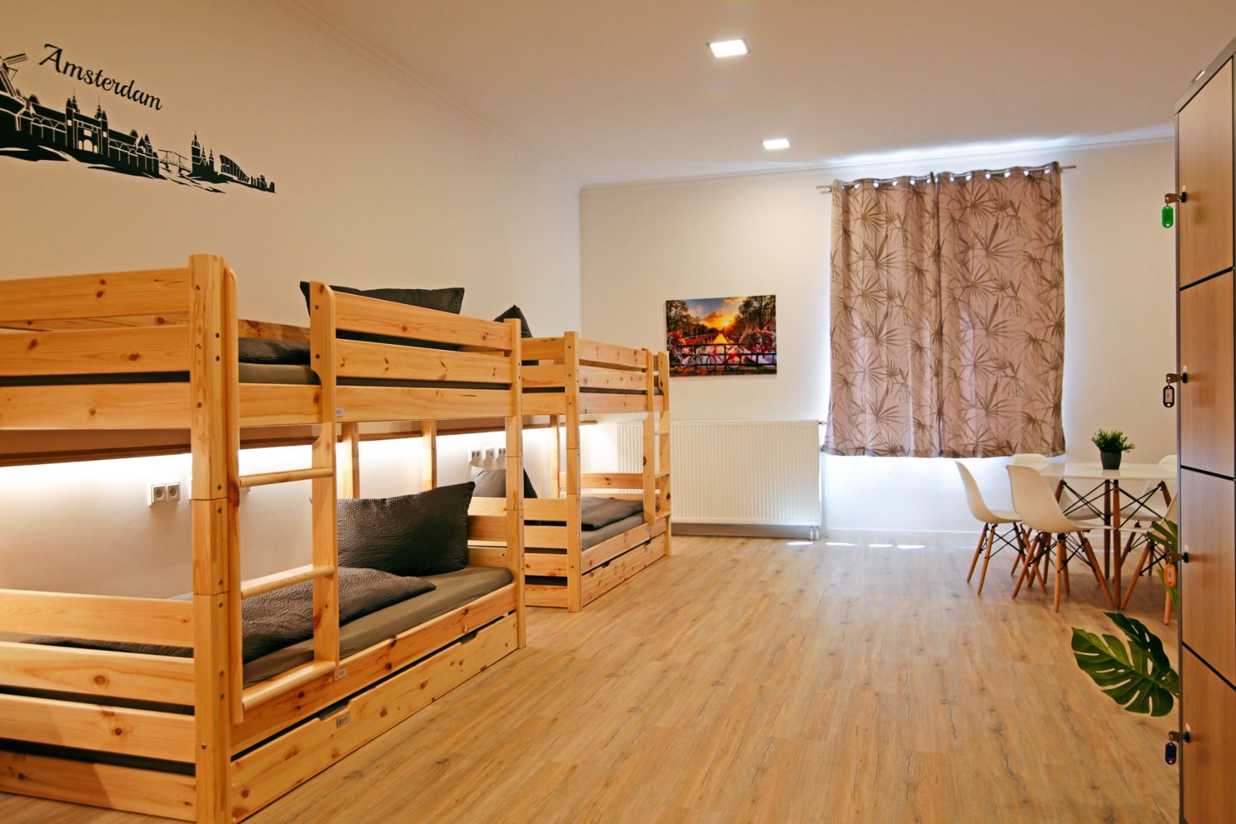 Ein helles, ordentliches Herbergszimmer im Sportpark Fun4You verfügt über Etagenbetten aus Holz, graue Bettwäsche, Spinde, einen Tisch mit Stühlen, ein Fenster mit gemusterten Vorhängen und Amsterdamer Wandkunst - ideal für Teambuilding in der Nähe des Id