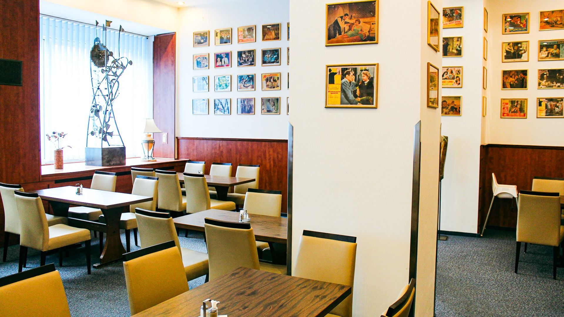 Das hell erleuchtete Restaurant im Hotel Marttel*** verfügt über hellbraune Stühle, dunkle Holztische, gerahmte Fotos und Kunstwerke sowie eine Metallskulptur und eine Lampe an einem großen Fenster mit vertikalen Jalousien in der Nähe der Grenznähe Deutsc