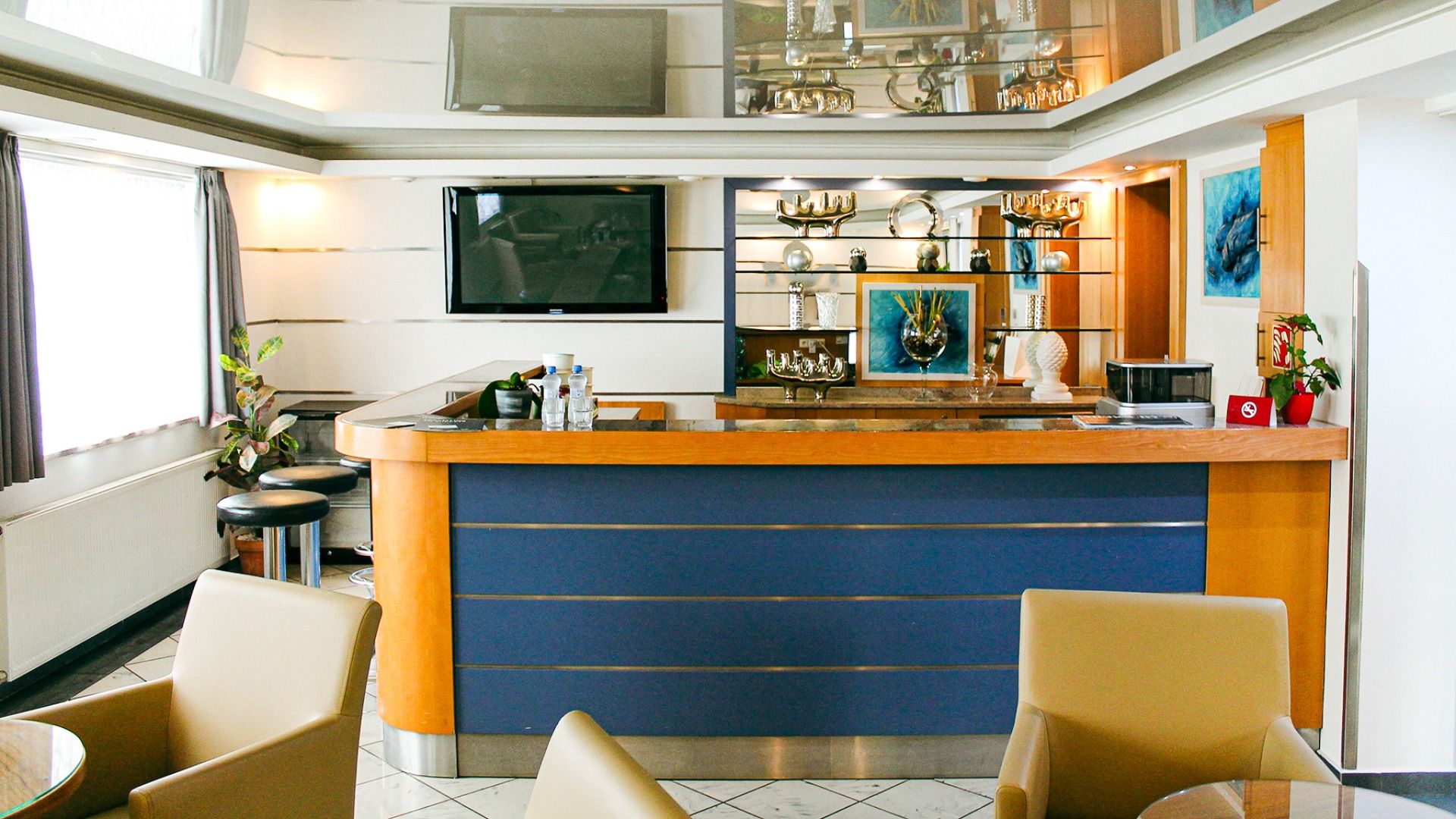 Das Hotel Marttel*** verfügt über eine moderne Lobby-Bar mit blauem und hölzernem Tresen, Glasvitrinen, Kunst, Flachbildfernseher, beigen Stühlen und Hockern an großen, mit Vorhängen versehenen Fenstern - ideal gelegen in Grenznähe zu Deutschland.