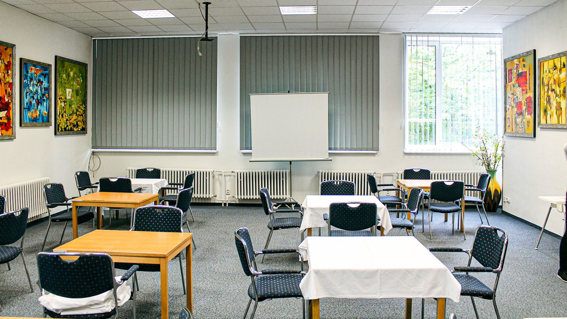 In einem hell erleuchteten Klassenzimmer im Hotel Marttel*** in der Nähe der Grenznähe zu Deutschland stehen Tische und Stühle, teilweise mit weißen Tischdecken, eine Projektionsfläche, große Fenster mit Jalousien und bunte Wandmalereien.