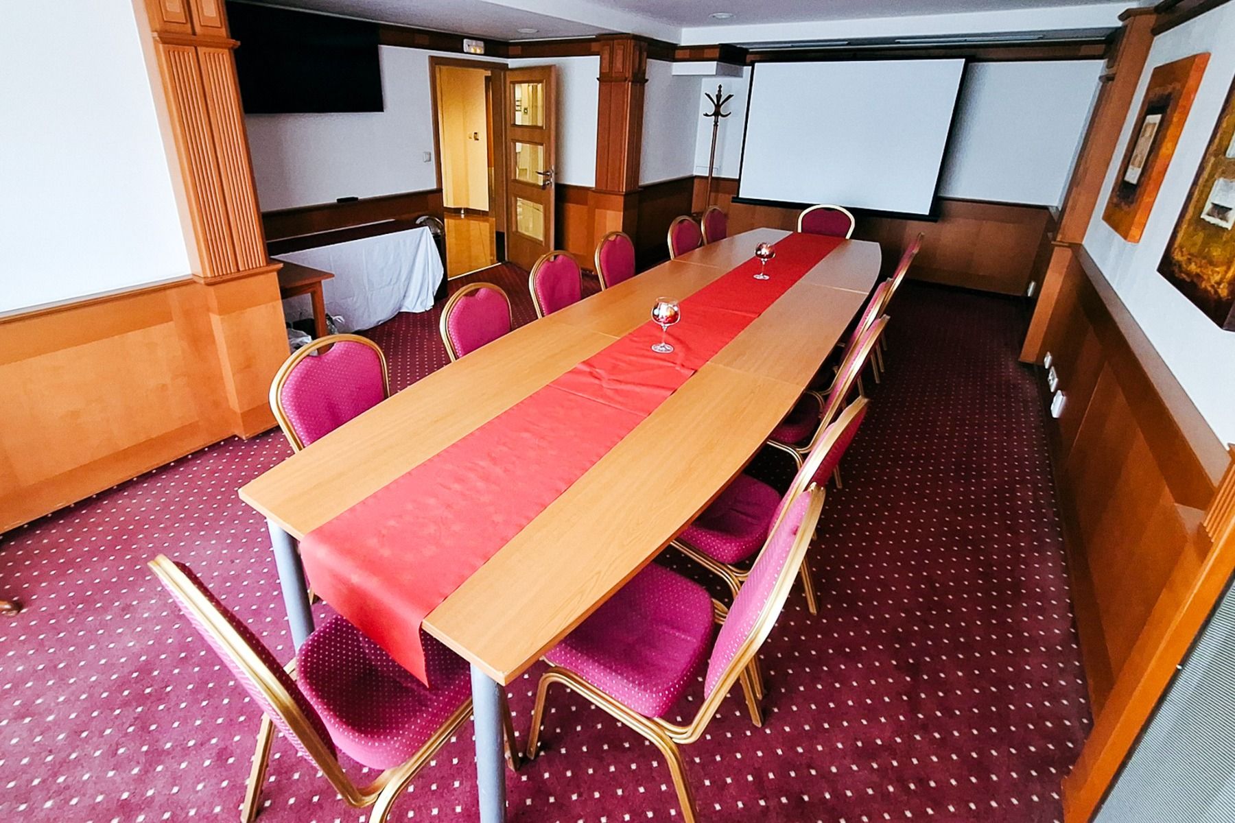 Ein Konferenzraum im Hotel Primavera mit einem langen, mit einem roten Läufer bedeckten Tisch, roten Stühlen, einem Projektionsschirm, einem Fernseher, Wandkunst und einem gemusterten roten Teppich.