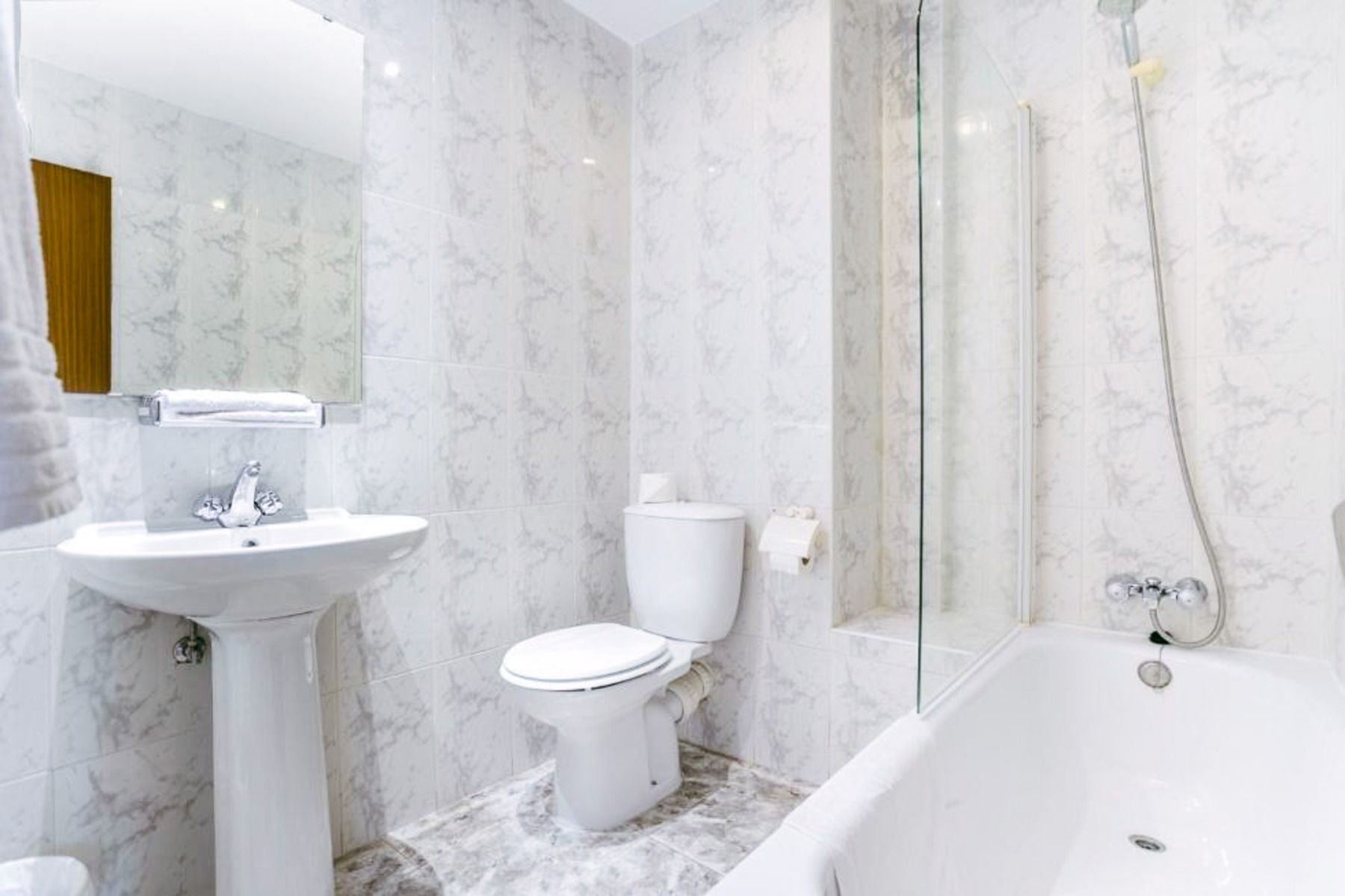 Das Hotel Torremolinos Centro verfügt über ein makelloses, weißes Badezimmer mit einem Waschbecken, einer Toilette und einer Badewanne mit Glasduschabtrennung, die mit Marmorfliesen ausgelegt ist. Die Handtücher liegen ordentlich auf einem Ständer für Sie