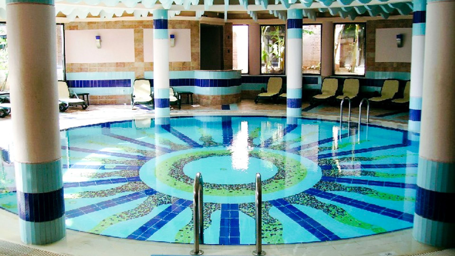 Limak Arcadia Hotel har et sirkulært innendørsbasseng med blå og grønne mosaikkfliser, solsenger, søyler og et tak dekorert med elegante mønstre og dempet belysning.