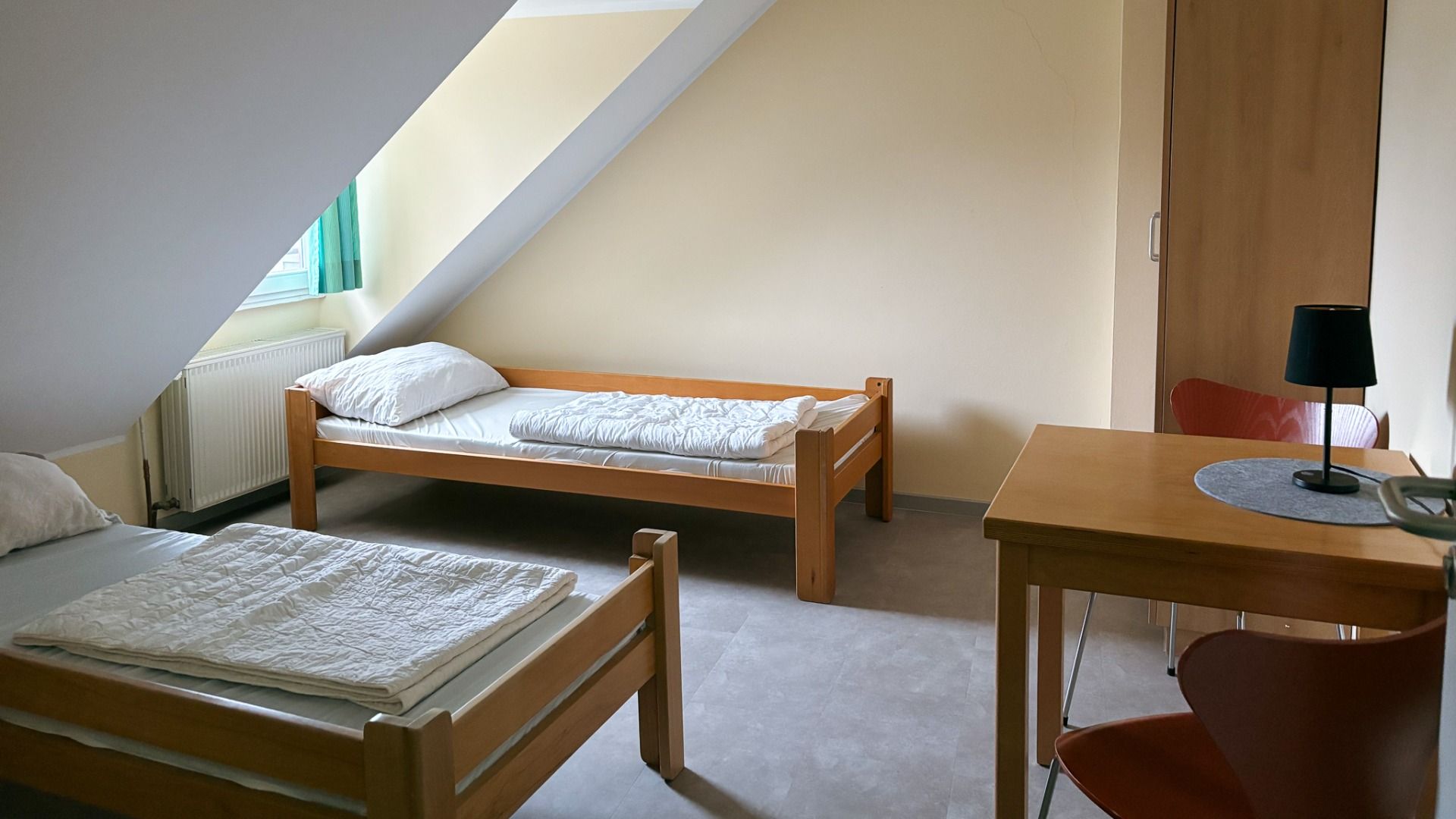 Das Gästehaus Nettersheim bietet ein einfaches, ordentliches Zimmer mit zwei Einzelbetten aus Holz, einem Schreibtisch mit Lampe und Stuhl, einem Fenster mit grünen Vorhängen für natürliches Licht, hellgelben Wänden, grauen Böden und einer funktionellen E