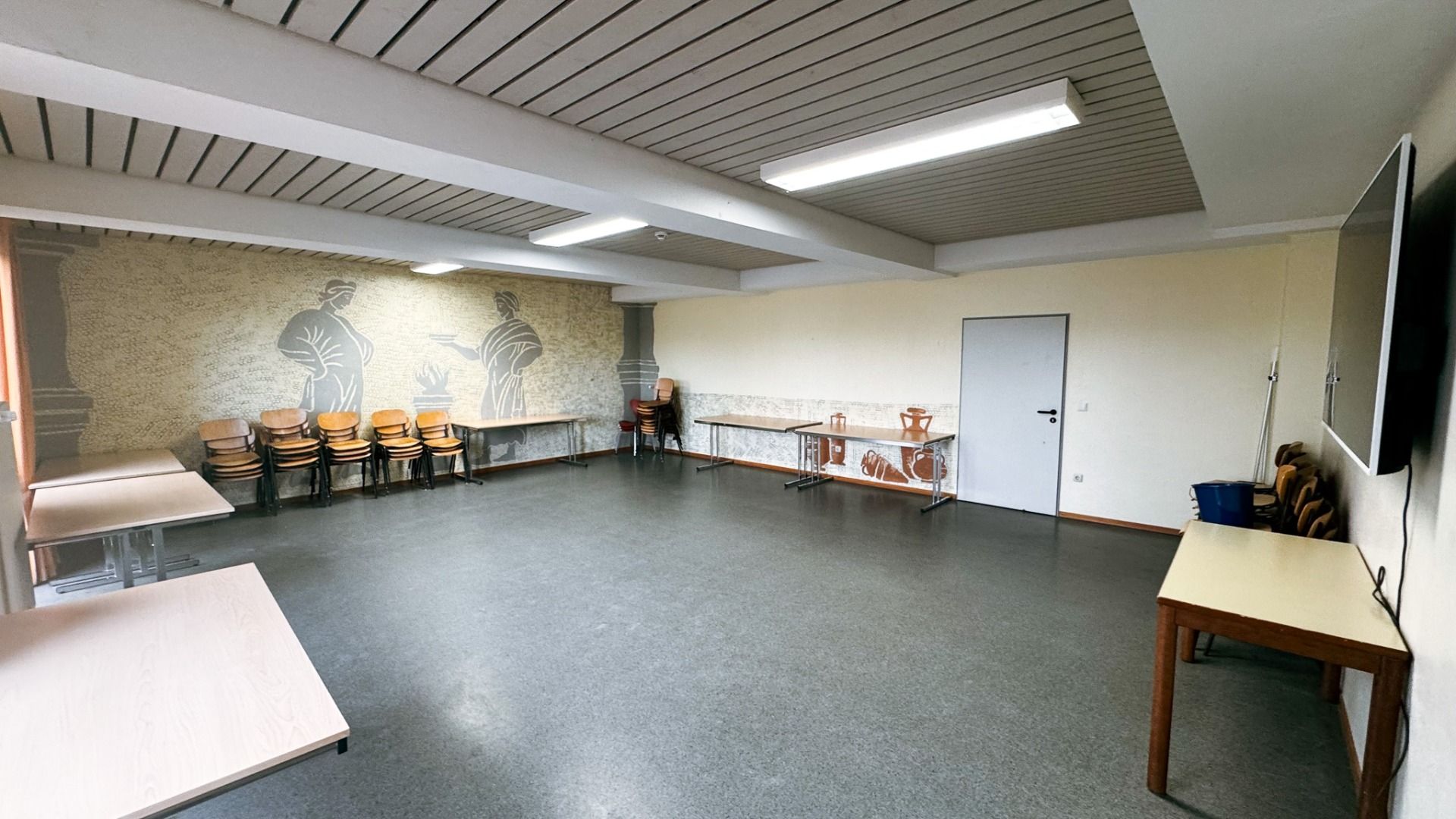 Ein geräumiges, leeres Klassenzimmer im Gästehaus Nettersheim ist mit einem Wandgemälde mit zwei Figuren, gestapelten Stühlen und Tischen, einer Tür, einem Flachbildfernseher, funksjonellem Fasiliteter, grauem Bodenbelag und Leuchtstofflampen ausgestattet