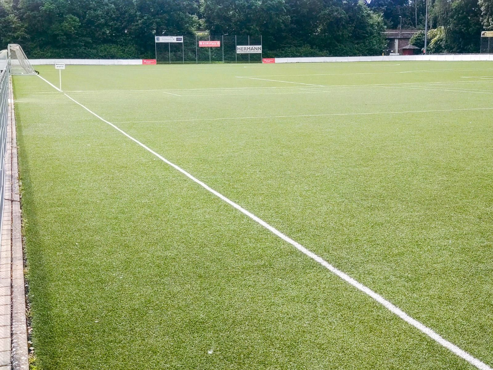 Ein Fußballplatz mit grünem Rasen und weißen Linien, leeren Tribünen auf der linken Seite und Bäumen in der Nähe der Sportanlage ist ideal für ein Trainingslager im Gästehaus Northeim. Im Hintergrund sind Schilder zu sehen.