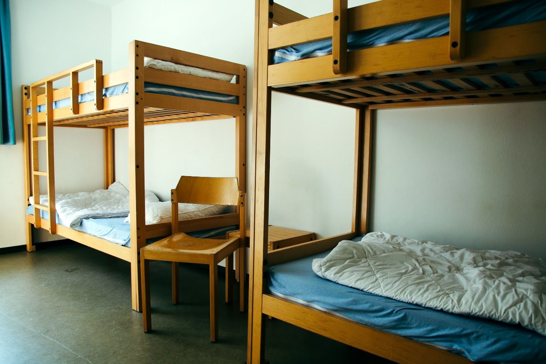 Ein praktischer Schlafsaal im Gästehaus Northeim besteht aus zwei hölzernen Etagenbetten mit blauen Matratzen und weißem Bettzeug sowie einem Holzstuhl und einem kleinen Tisch zwischen den Betten. Durch ein Fenster auf der linken Seite strömt Sonnenlicht 