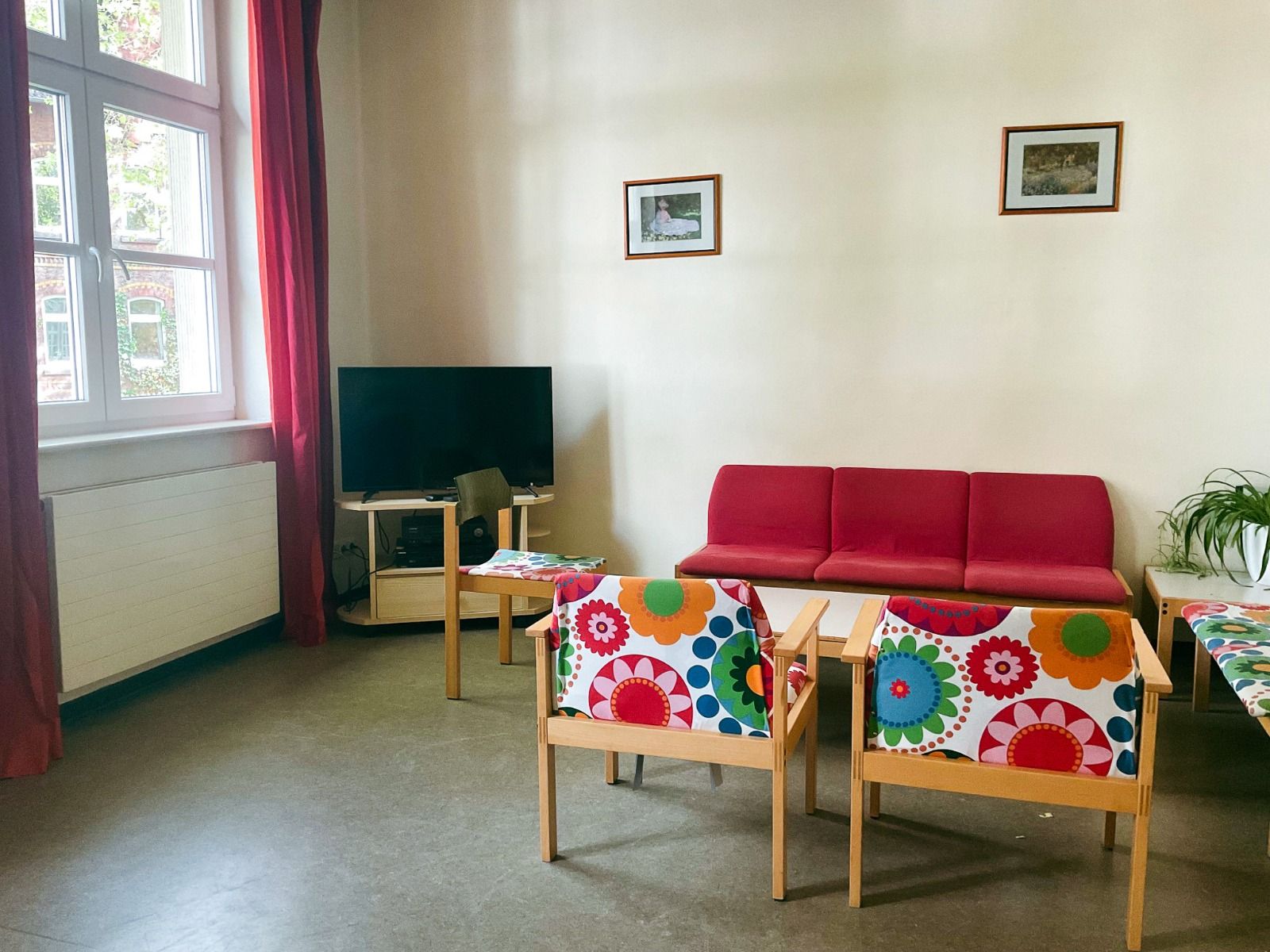 Ein helles Zimmer im Gästehaus Northeim verfügt über ein großes Fenster, rote Vorhänge, ein rotes Sofa und bunte geblümte Stühle - ideal zum Entspannen nach dem Besuch der nahe gelegenen Sportanlage. TV, gerahmte Bilder und eine Grünpflanze sorgen für Gem