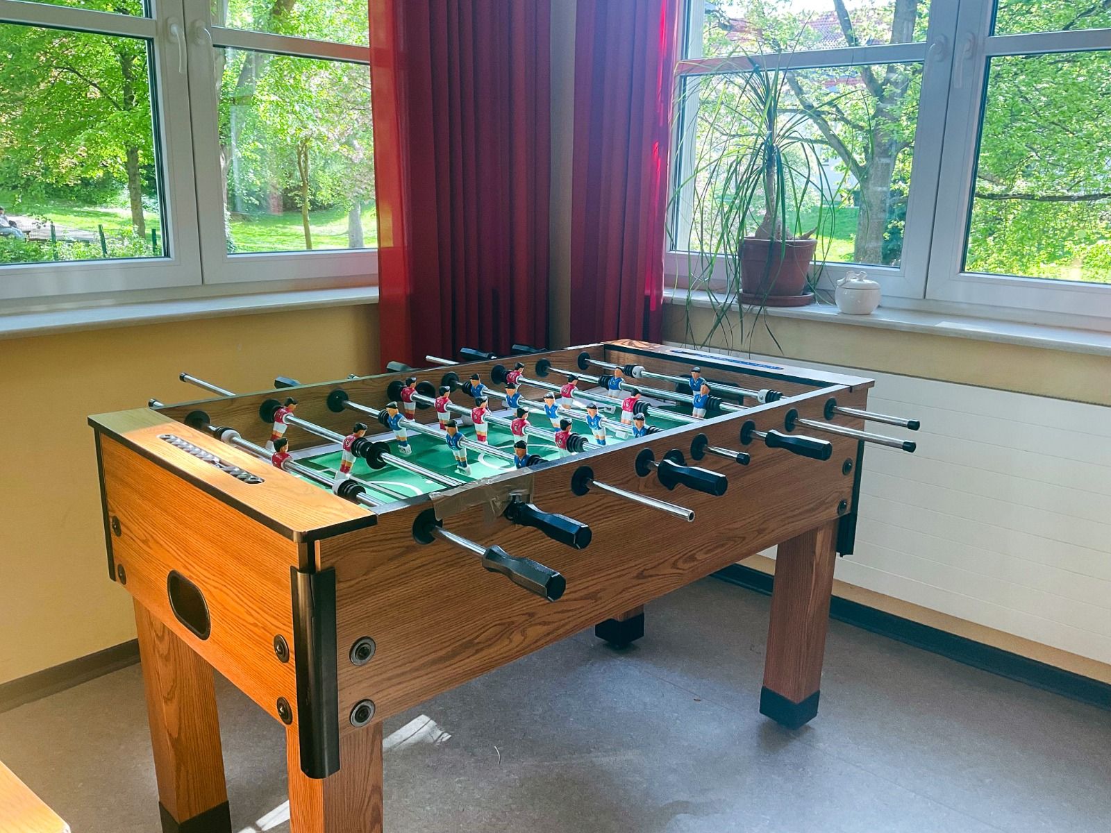 Das Gästehaus Northeim verfügt über einen Kickertisch mit schwarzen Griffen in einem sonnenbeschienenen Raum mit großen Fenstern, roten Vorhängen und Grünpflanzen - ideal für ein Trainingslager oder als sportfreundliche Unterkunft mit Blick auf die üppige