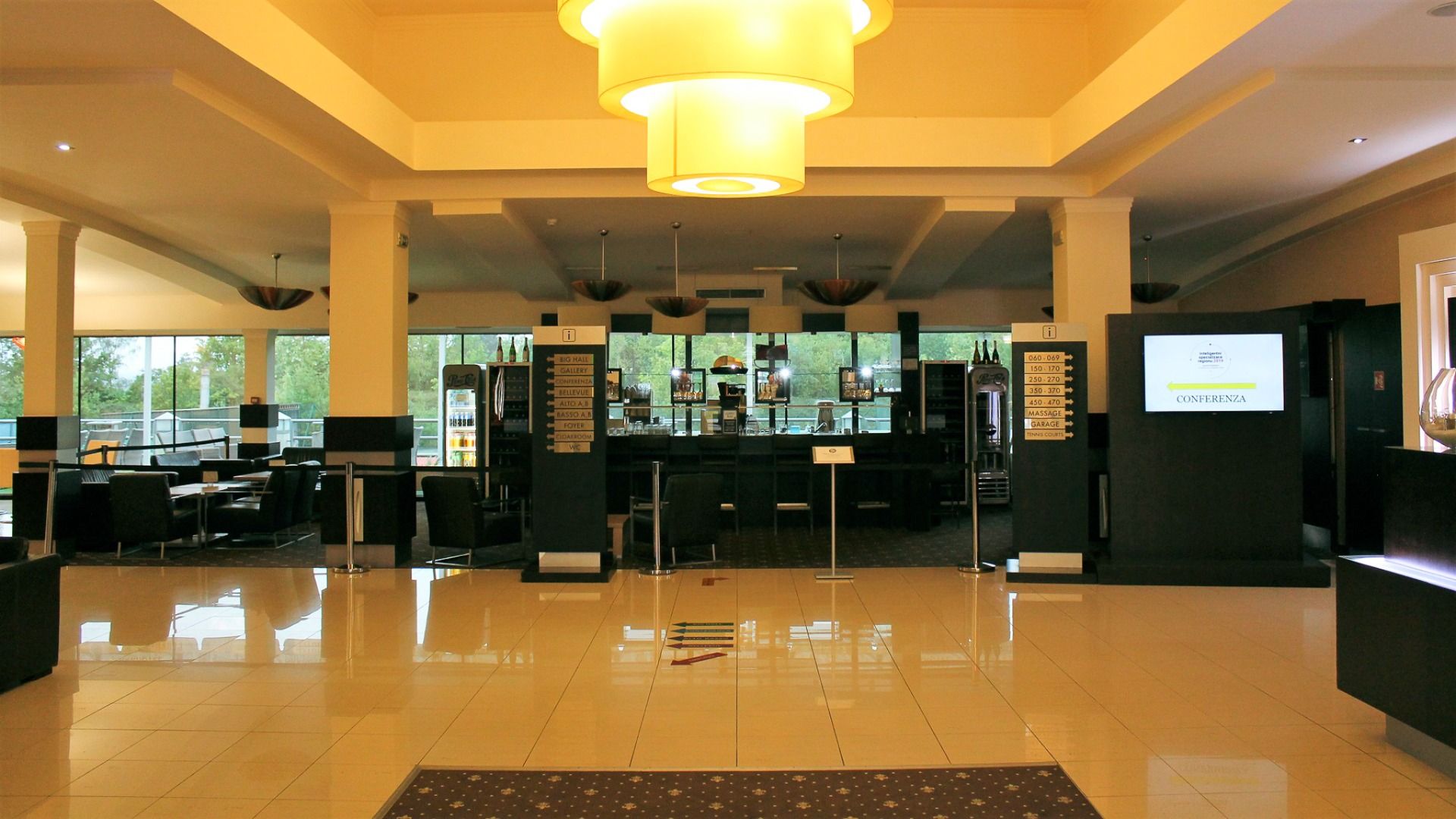 Die geräumige Lobby des Parkhotels in Pilsen bietet glänzende Fliesenböden, moderne Sitzgelegenheiten, eine Bar und eine große Deckenleuchte. Auf einem digitalen Schild steht CONFERENCE. Große Fenster lassen das Stadtzentrum Pilsen mit natürlichem Licht e