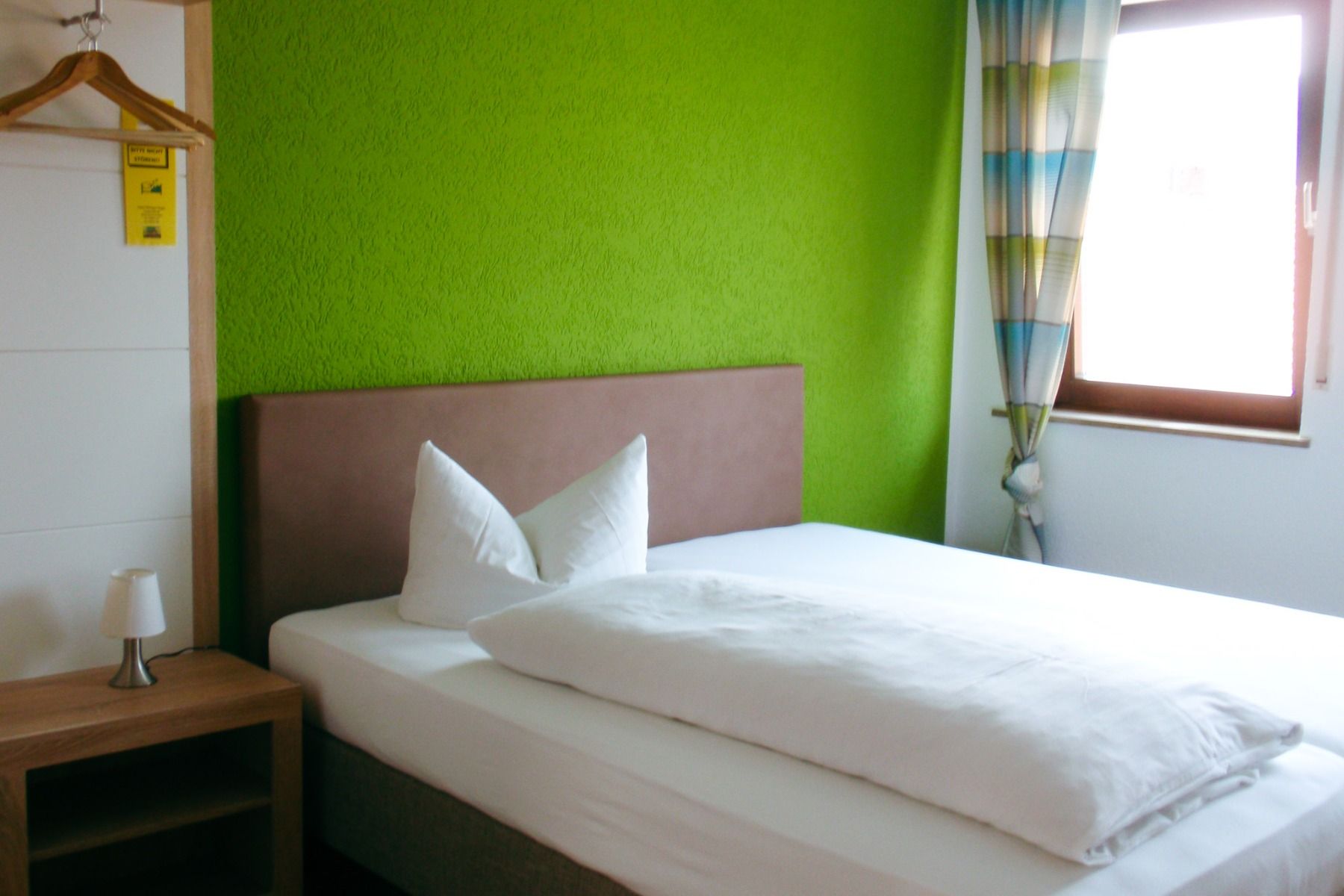 Ein ordentlich gemachtes Bett mit weißem Bettzeug im Weingut Hotel Goger steht an einer grünen Wand. Ein hölzerner Nachttisch mit einer Lampe und ein offenes Fenster mit gestreiften Vorhängen bringen Licht ins Zimmer, während Kleiderbügel mit einem Schild