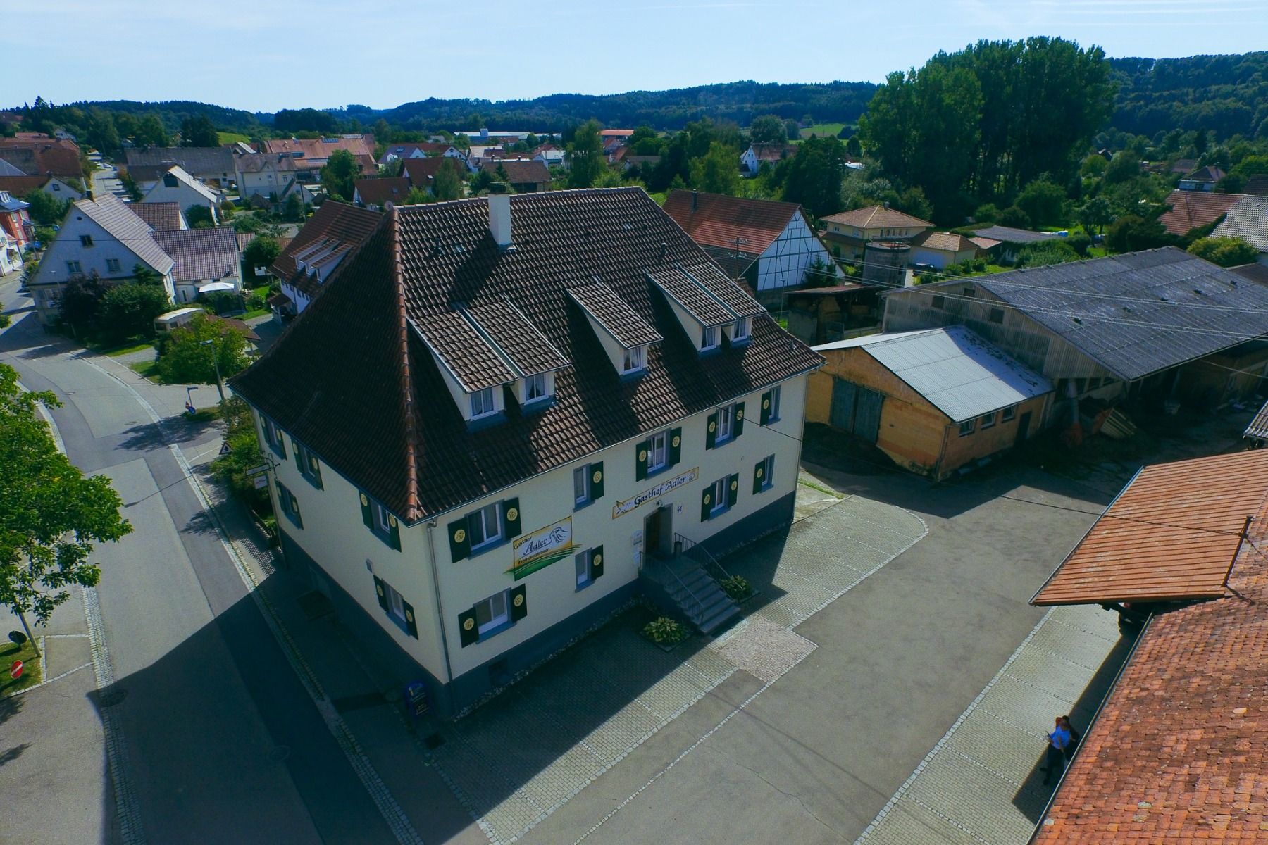Luftaufnahme des Gasthofs Adler, eines geräumigen weißen Gebäudes mit braunem Ziegeldach und grünen Fensterläden, das mit einem Fassadenschild versehen ist und sich in einer ruhigen Umgebung zwischen benachbarten Häusern in einem kleinen Dorf befindet.