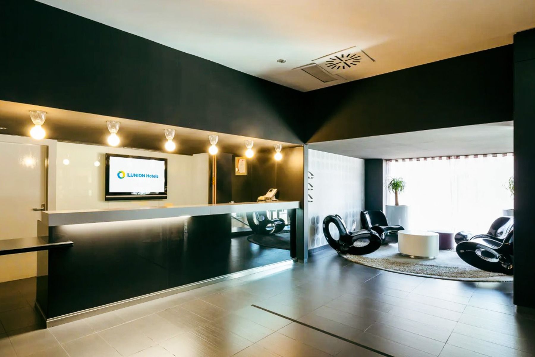 Moderne hotellresepsjon med svart skranke, overlys og en TV som viser Hotel ILUNION Bel Art Barcelona. Til høyre er det sitteplasser med utsikt over byens berømte Fußballkultur Barcelona gjennom et stort vindu.