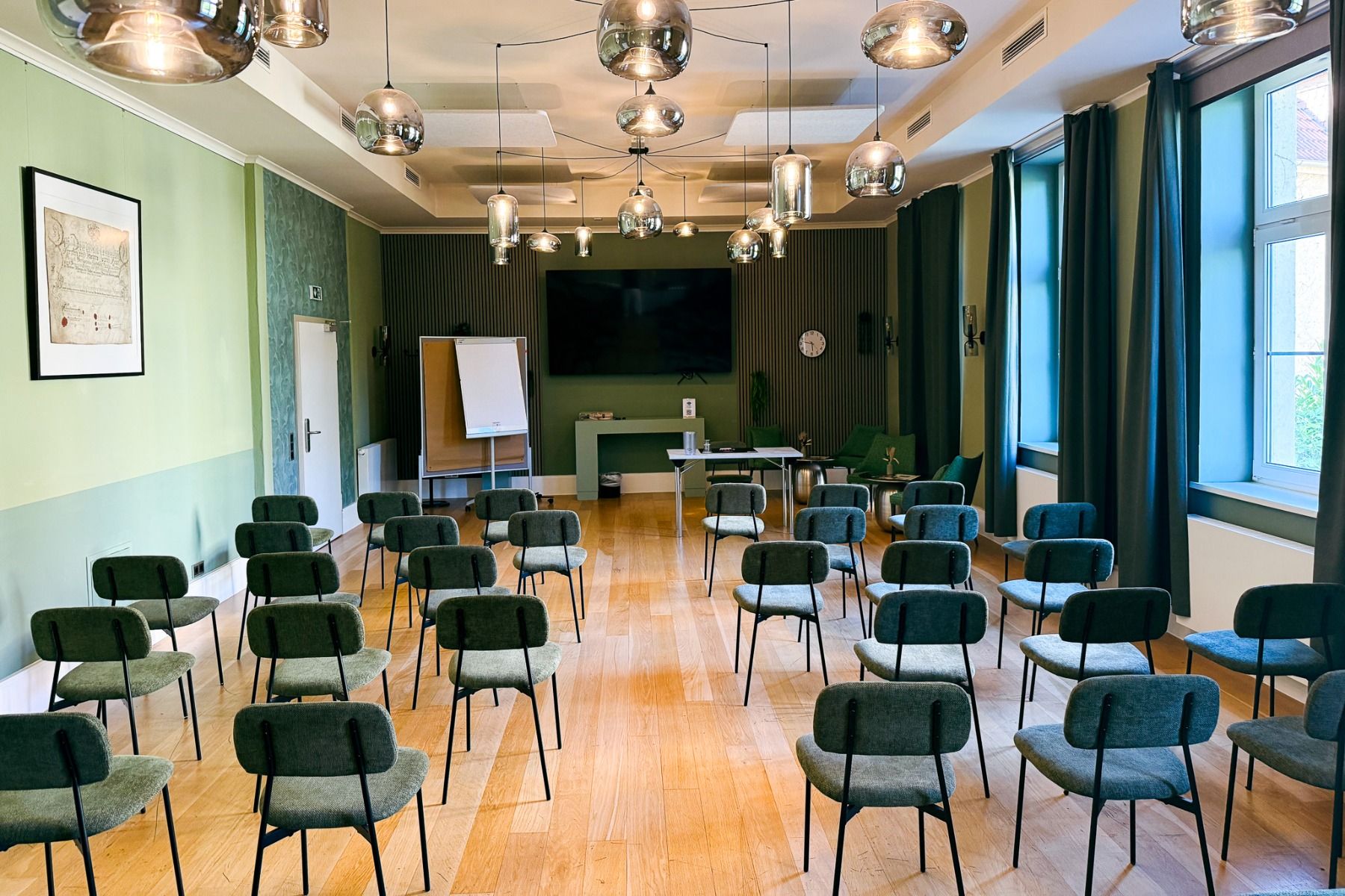 Et lyst, moderne konferanserom med rekker av grønne stoler vendt mot en skjerm, en tavle og bord foran i rommet. Pendellamper henger fra taket, og store vinduer slipper inn naturlig lys – alt dette i Hotel Dorotheenhof, der moderne design møter historisk