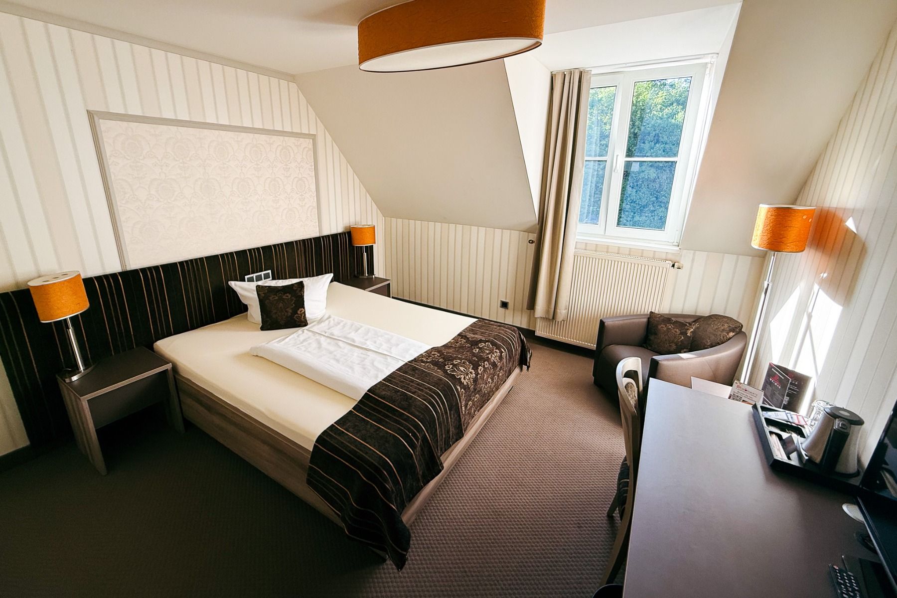 Et ryddig rom på Hotel Dorotheenhof har en dobbeltseng, to lamper, et romslig vindu, en lenestol, et skrivebord og en innredning i nøytrale beige- og brune farger – noe som gjør det til det perfekte tilfluktsstedet både for fritidsopphold og fotballtrenin