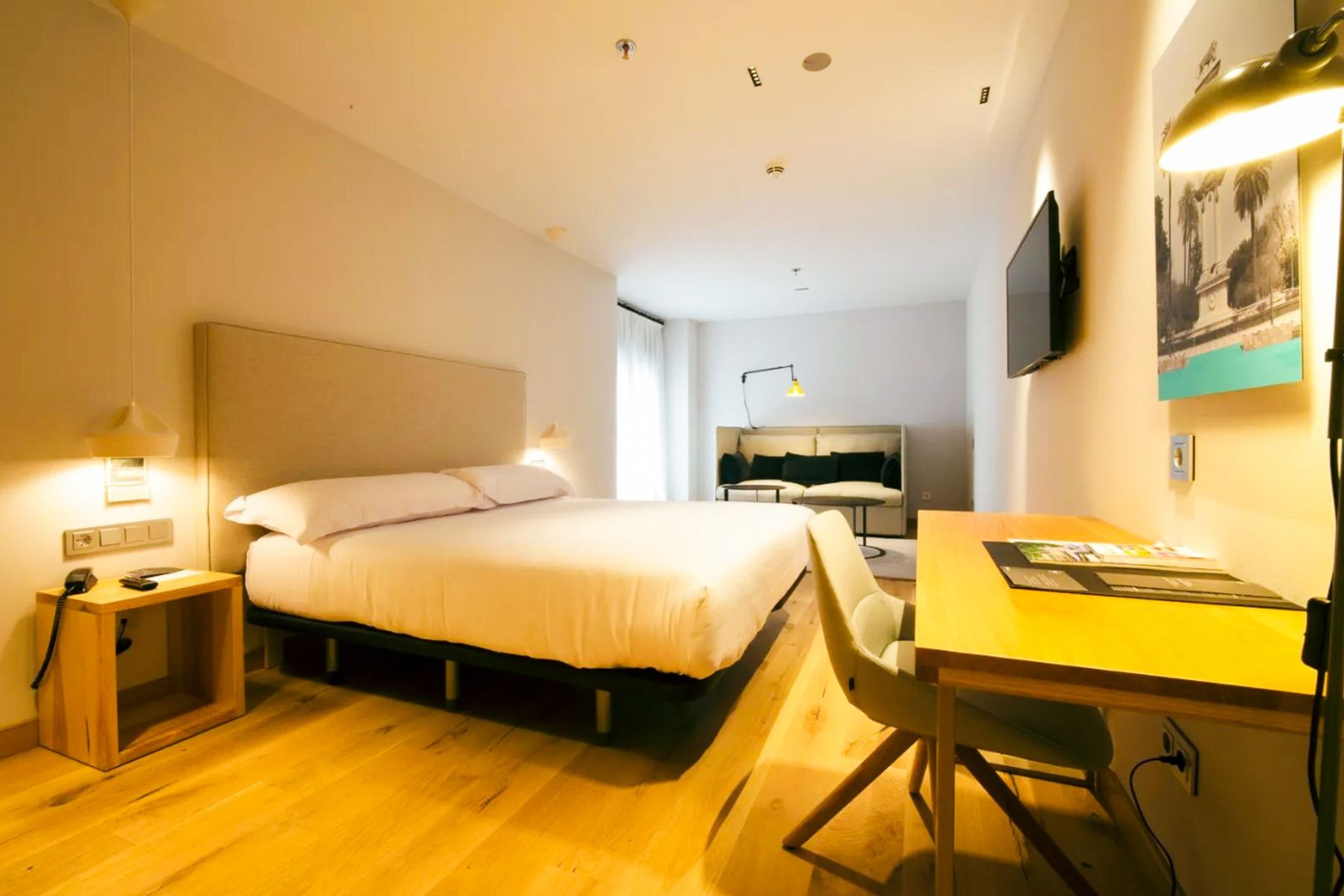Et moderne rom på Hotel Zenit Sevilla har en stor seng, tregulv, nattbord, skrivebord med stol og lampe, veggmontert TV, sofa ved vinduet og dempet belysning - perfekt for Fußball-Sportgruppen på Hotel Zenit Sevilla.