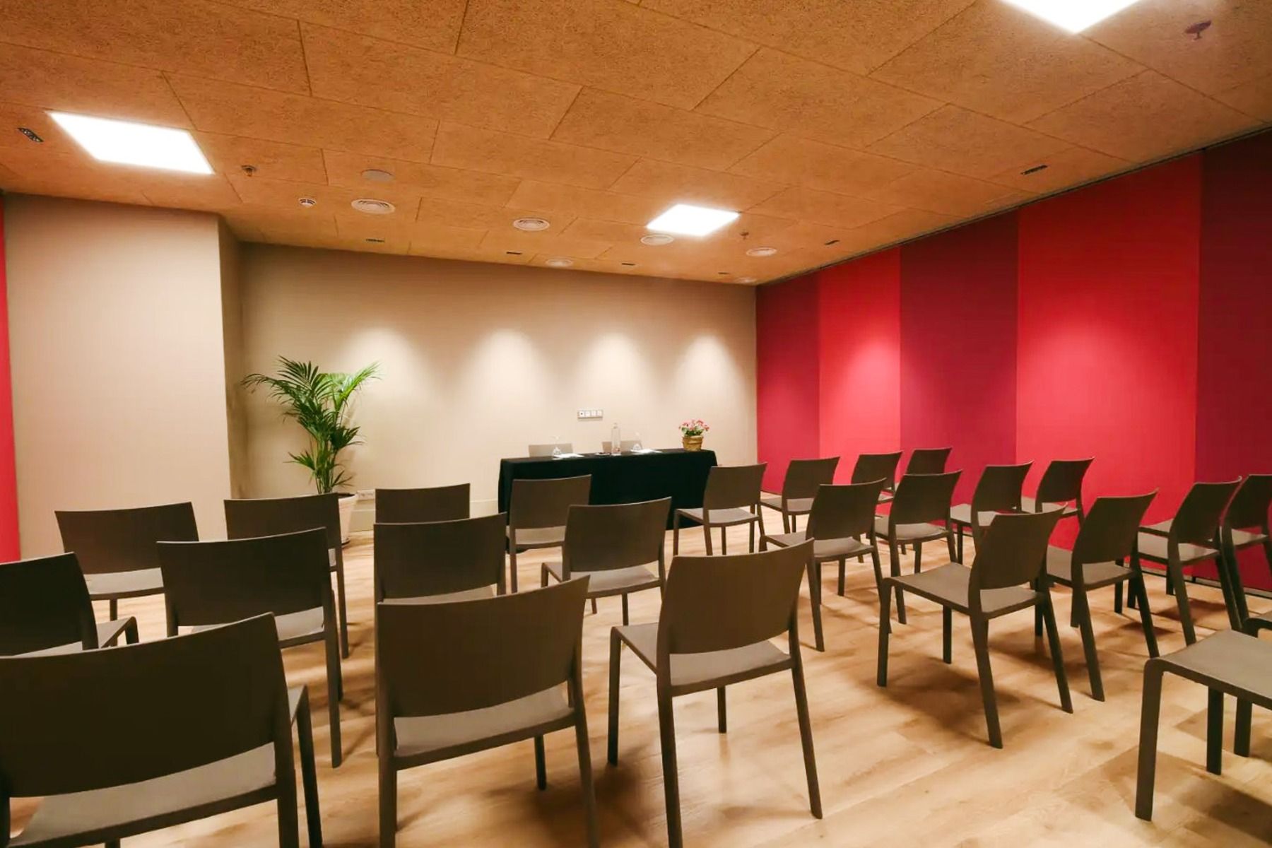 I et lite konferanserom på Hotel Zenit Sevilla står rader med grå stoler foran et bord med planter og blomster. Røde og beige vegger, tregulv og taklamper skaper en ideell ramme for møter i Fotball-Sportgruppen.