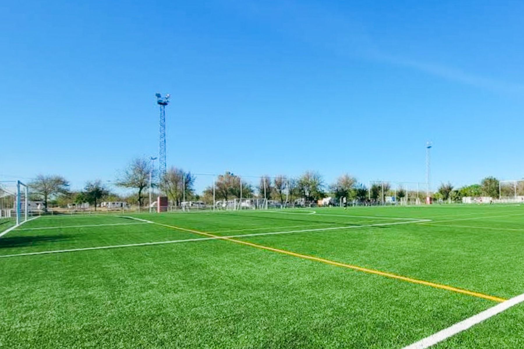 En klar, solrik dag på en tom fotballbane med lysegrønt kunstgress - ideelt for Fußball-Sportgruppen som bor på Hotel Zenit Sevilla. Scenen har hvite og gule markeringer, målstolper, trær og høye flomlys.