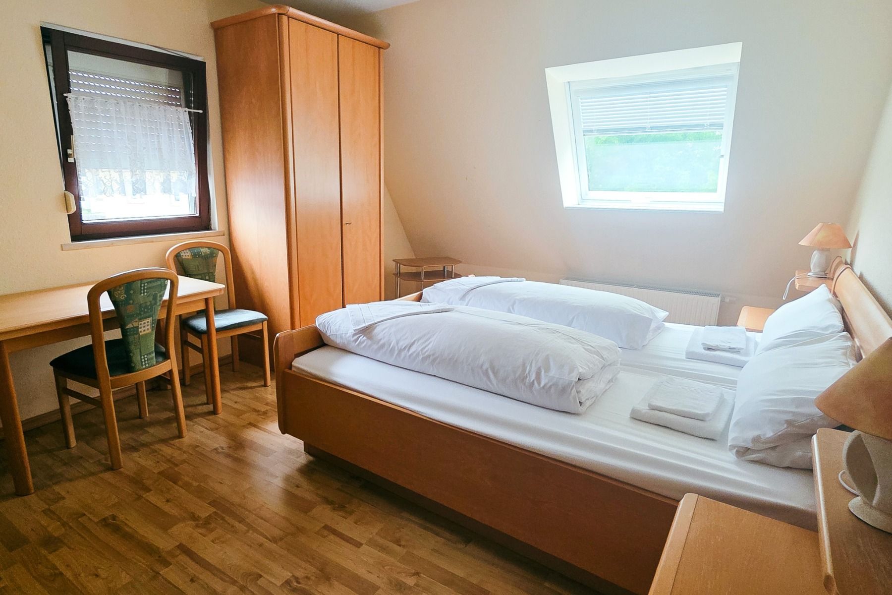 Et lyst, enkelt rom på Gasthof Kapellenhof i Nürnberg har en dobbeltseng, tremøbler, garderobe, skrivebord med to stoler ved vinduet og sollys som strømmer inn gjennom et skrått takvindu.