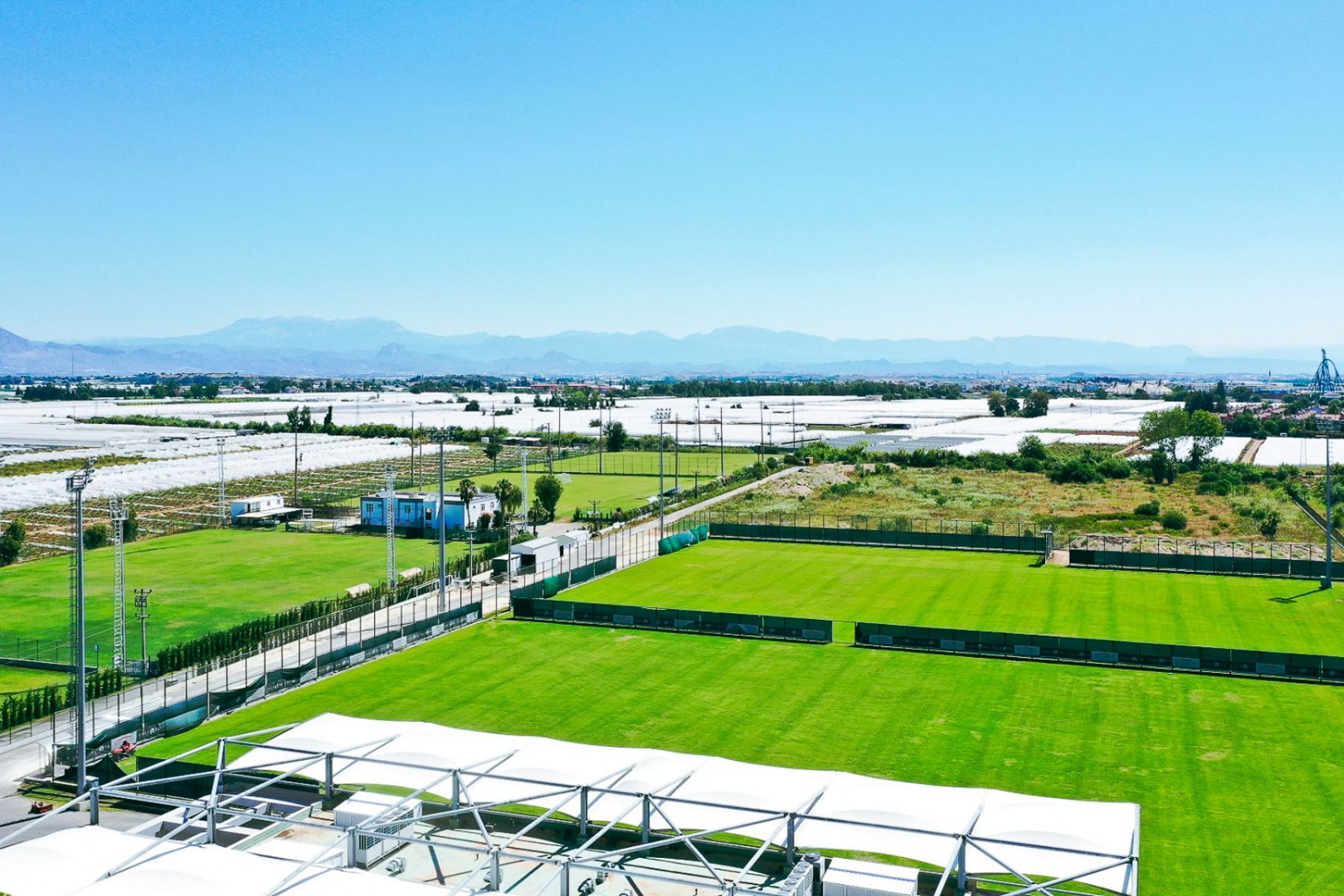 Flyfoto av flere grønne fotballbaner i nærheten av Limak Arcadia-hotellet, ideelt for en fotballtreningsleir i Tyrkia, omgitt av gjerder, åpne jorder, fjell i det fjerne og en klar blå himmel.