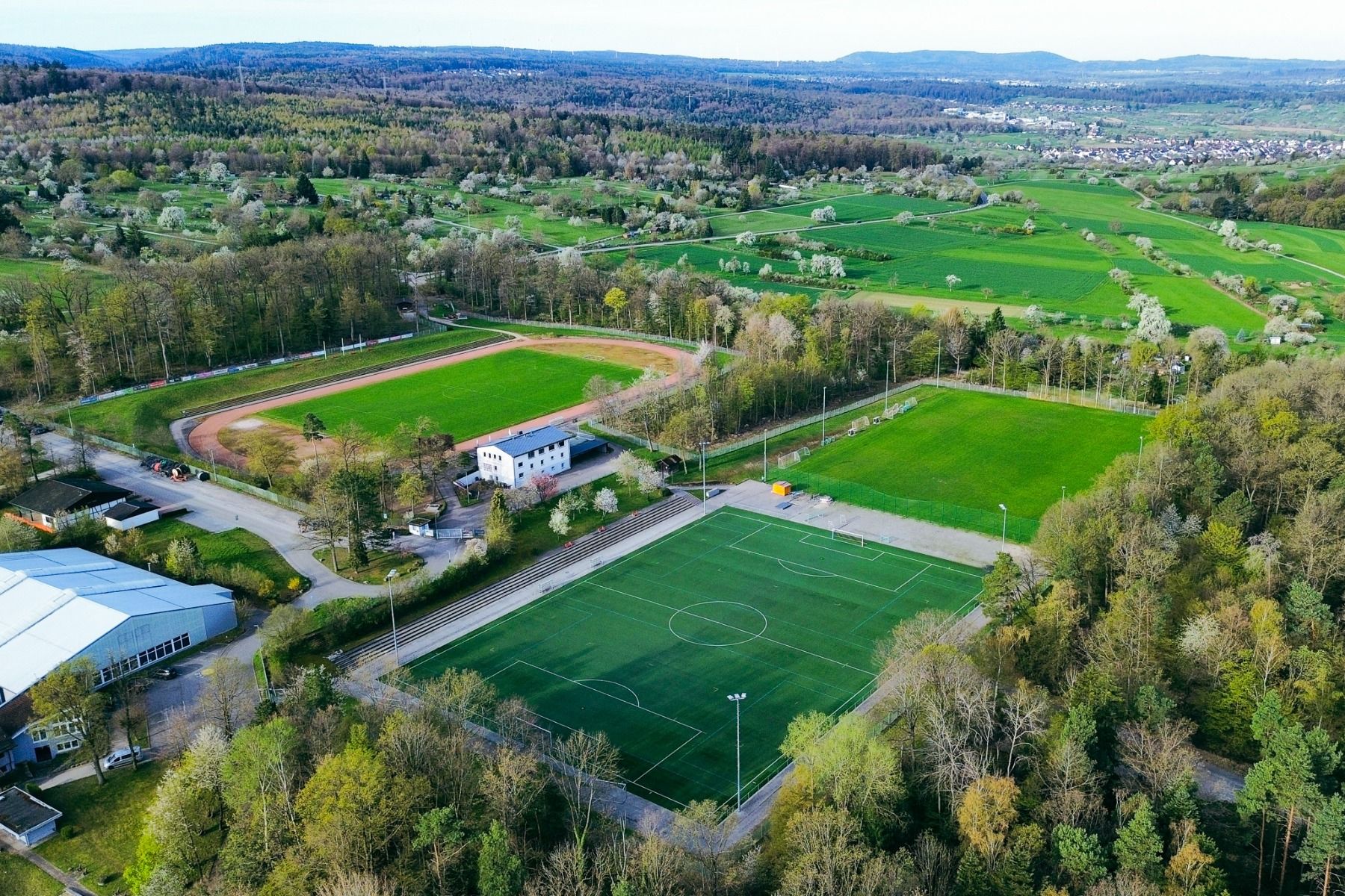 Luftfoto av et idrettsanlegg med en fotballbane med kunstgress, en friidrettsbane og flere grønne områder – ideelt for en treningsleir i Pforzheim med overnatting på Mercure Hotel Pforzheim – omgitt av trær og landlige omgivelser, med hus og åser i det fj