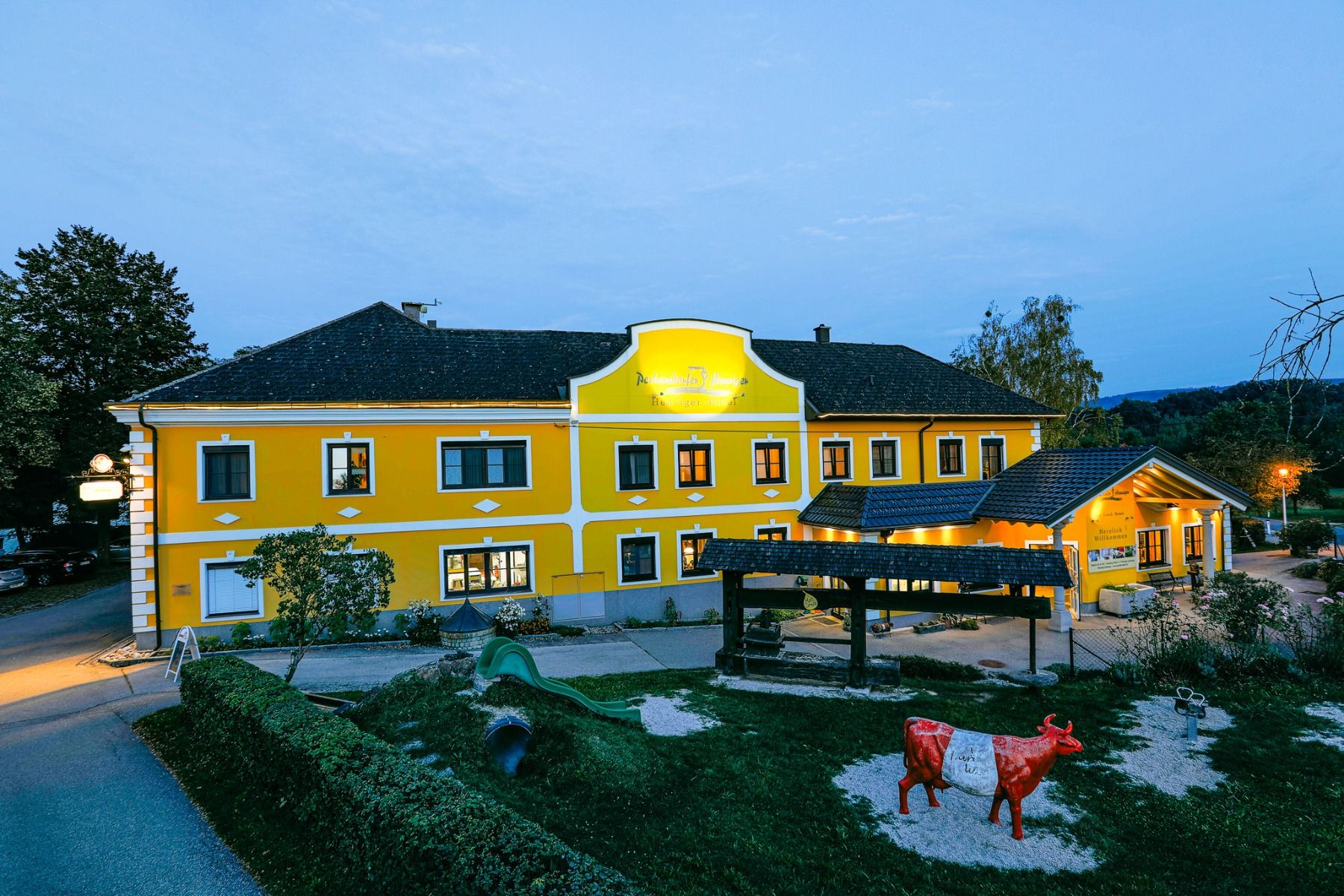 Hotellet Perbersdorfer Heuriger i Neuhofen an der Ybbs fremstår i et varmt lys i skumringen med sin knallgule fasade og hvite paneler. En rød og hvit kuskulptur hviler på grus omgitt av grøntområder nær den innbydende inngangen.