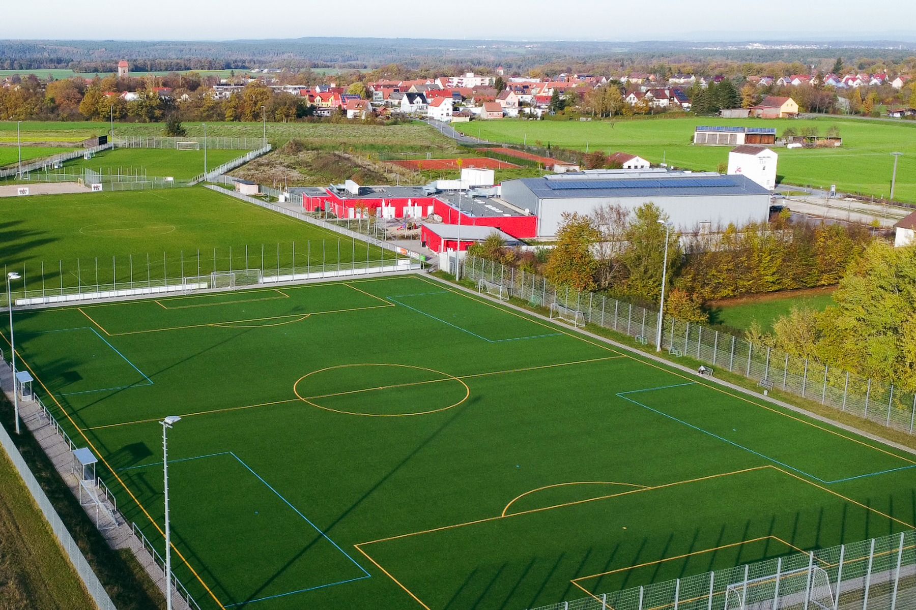 Flyfoto av en grønn fotballbane med gule og blå streker, omgitt av trær, gjerder, bygninger og jordbruksland - et steinkast fra Gasthof Kapellenhof, som er kjent for sin hjemmekoselige mat og koselige atmosfære i byen.