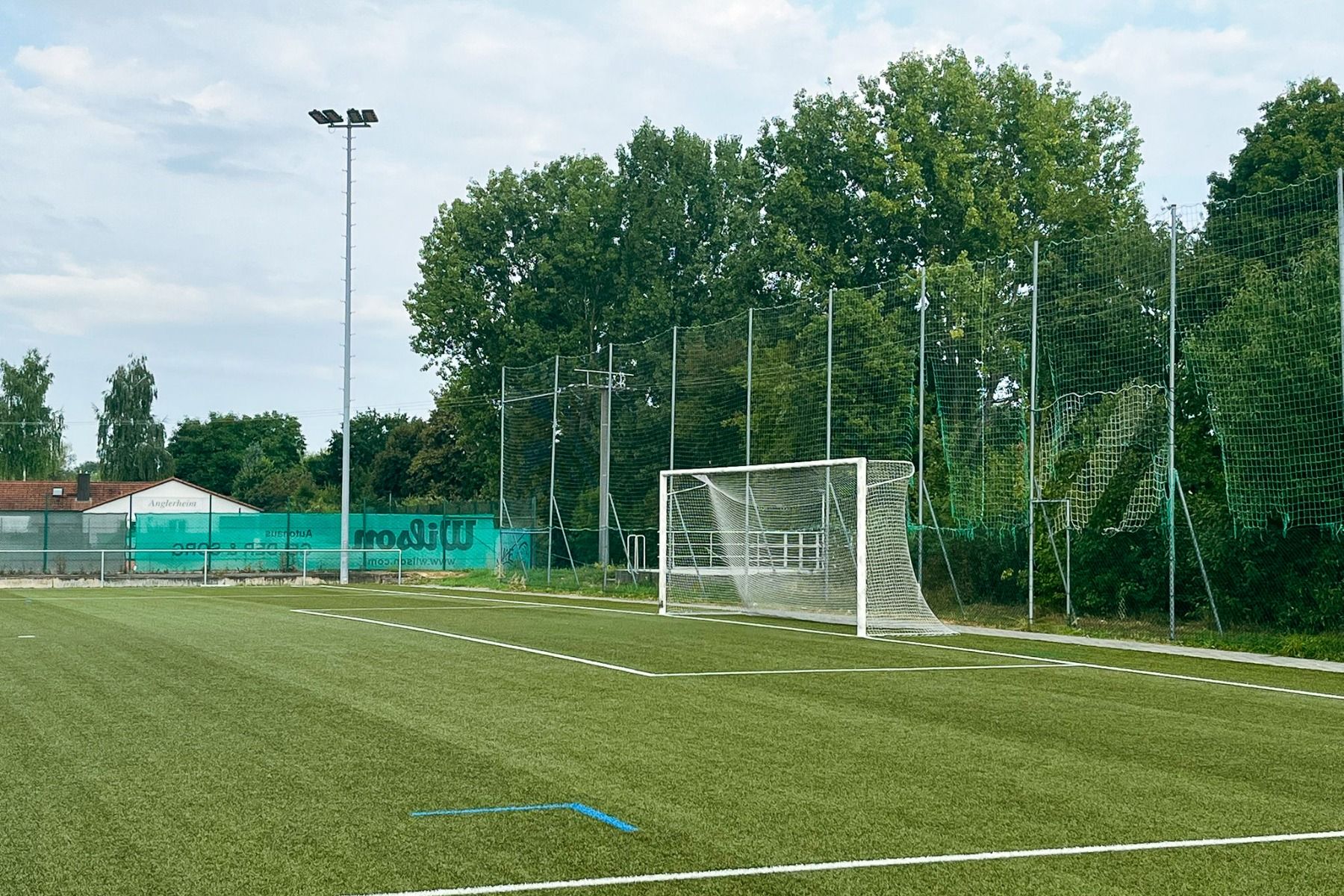 Ein Fußballtor steht auf einem leeren Kunstrasenplatz, der von hohen grünen Netzen und Bäumen begrenzt wird. Im Hintergrund ist das Weingut Hotel Goger unter einem teilweise bewölkten Himmel zu sehen.