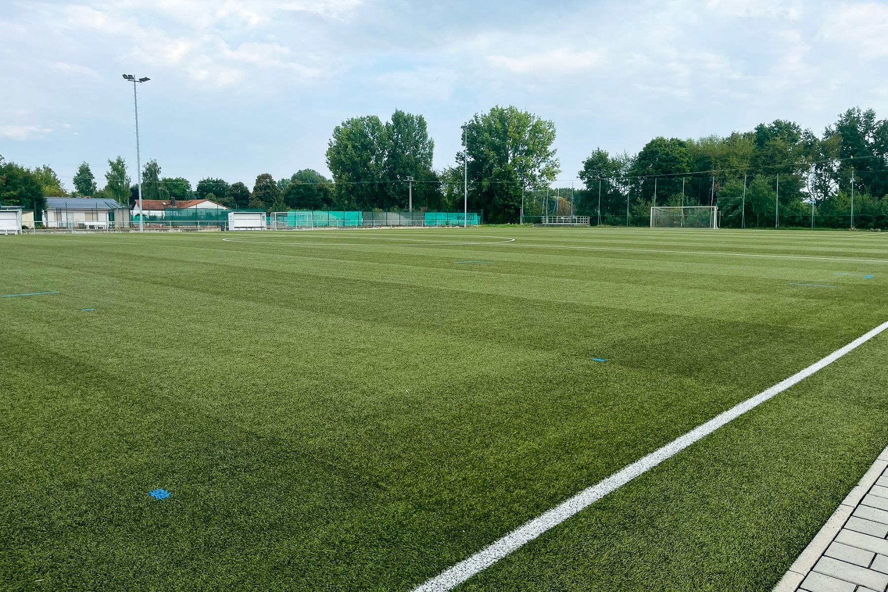 Weite Sicht auf einen leeren, gepflegten Fußballplatz mit grünem Kunstrasen und weißen Linien in der Nähe des Weingut Hotels Goger. Bäume, Gebäude und ein Weinberg erscheinen im Hintergrund unter einem teilweise bewölkten Himmel.