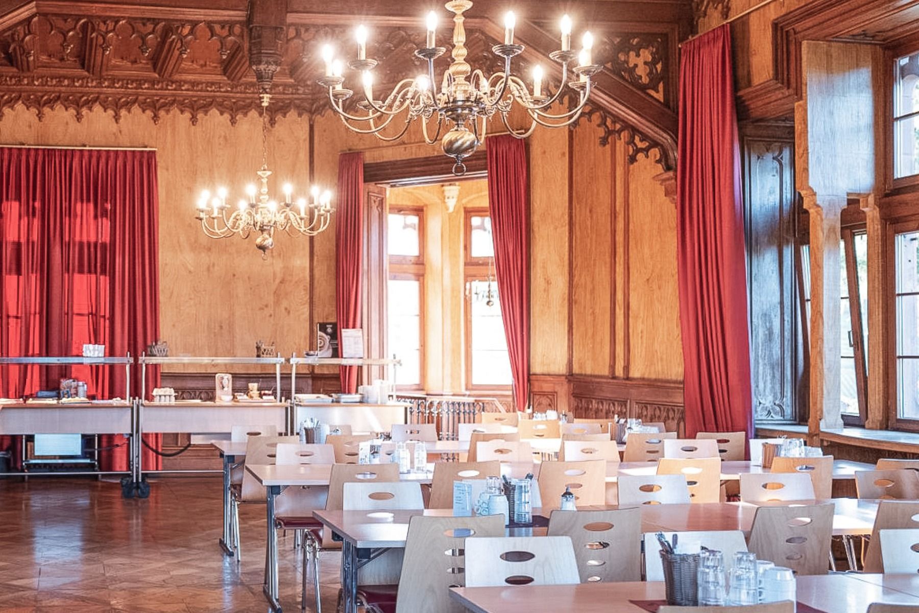 En elegant innredet spisesal med trevegger, lysekroner, røde gardiner og store vinduer. Den ligger i gjestehuset Schloss Ortenberg i hjertet av Schwarzwald. Bordene og stolene er pent ordnet og dekket med glassflasker, kopper og bestikk – et innbydende ro