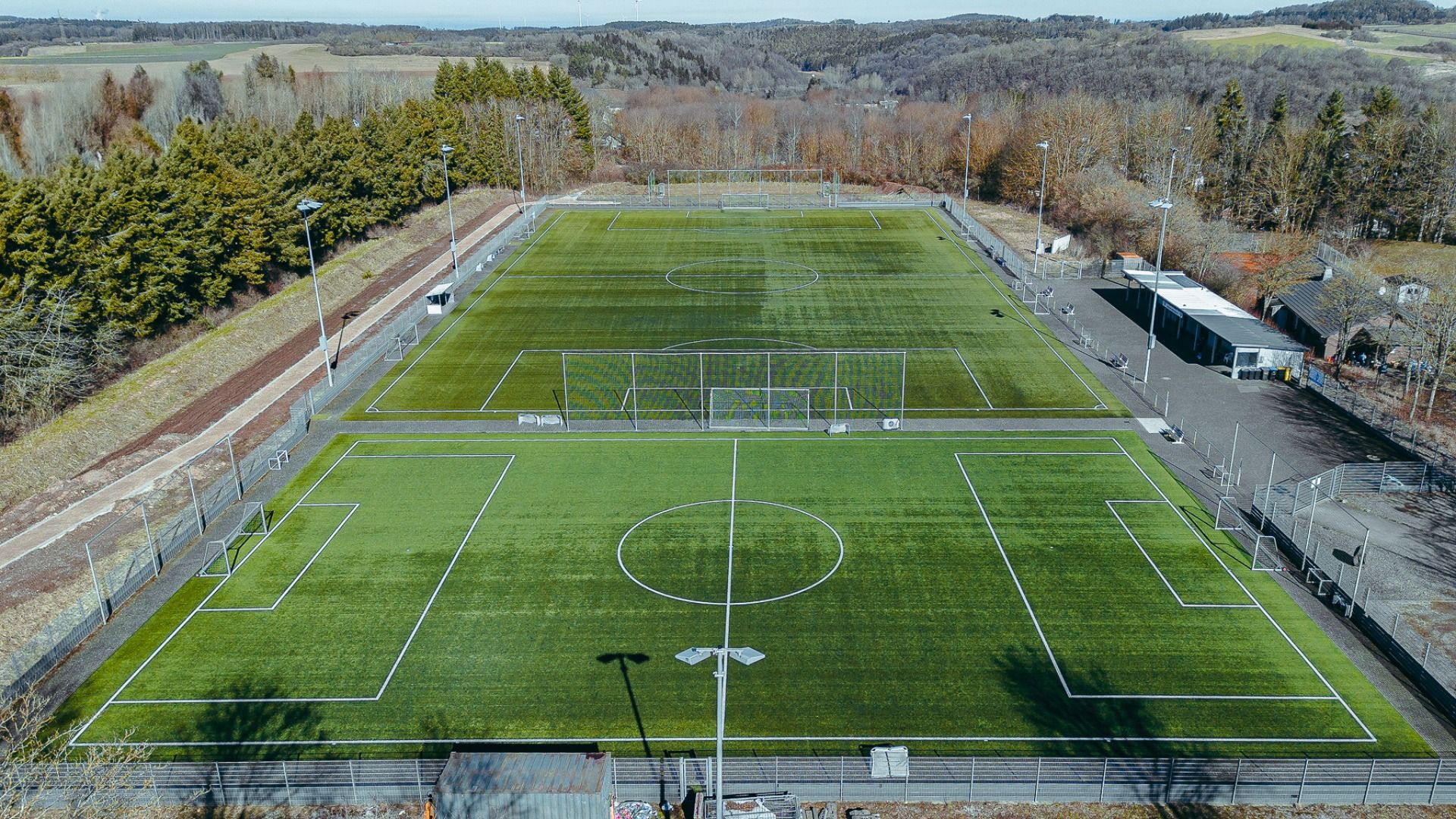 Luftaufnahme des Gästehauses Nettersheim: zwei nebeneinander liegende Kunstrasen-Fußballplätze mit funktionalen Einrichtungen, eingezäunt und eingebettet in malerische Wälder und Hügel an einem klaren Tag.