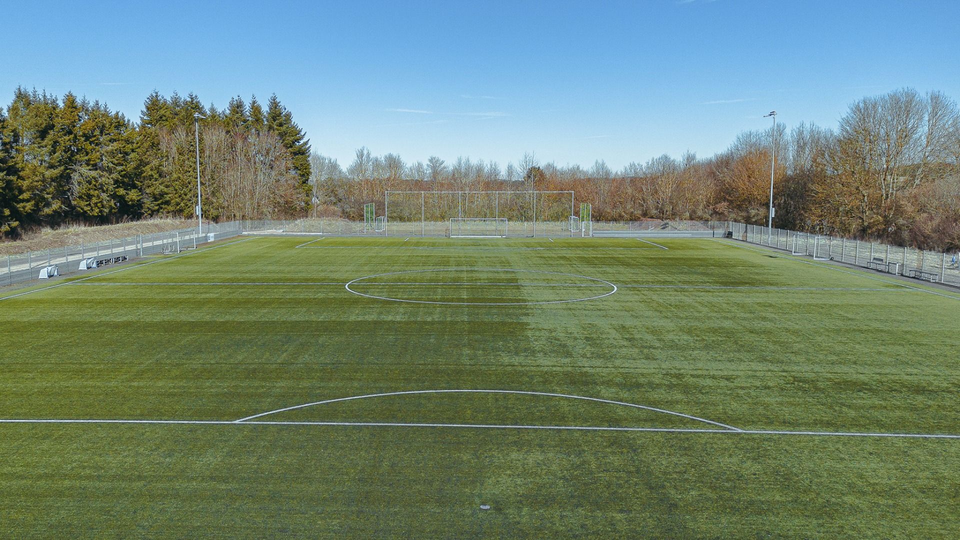 Ein breiter, leerer Fußballplatz mit Kunstrasen liegt neben dem Gästehaus Nettersheim, umgeben von einem Metallzaun und herbstlichen Bäumen unter einem strahlend blauen Himmel.