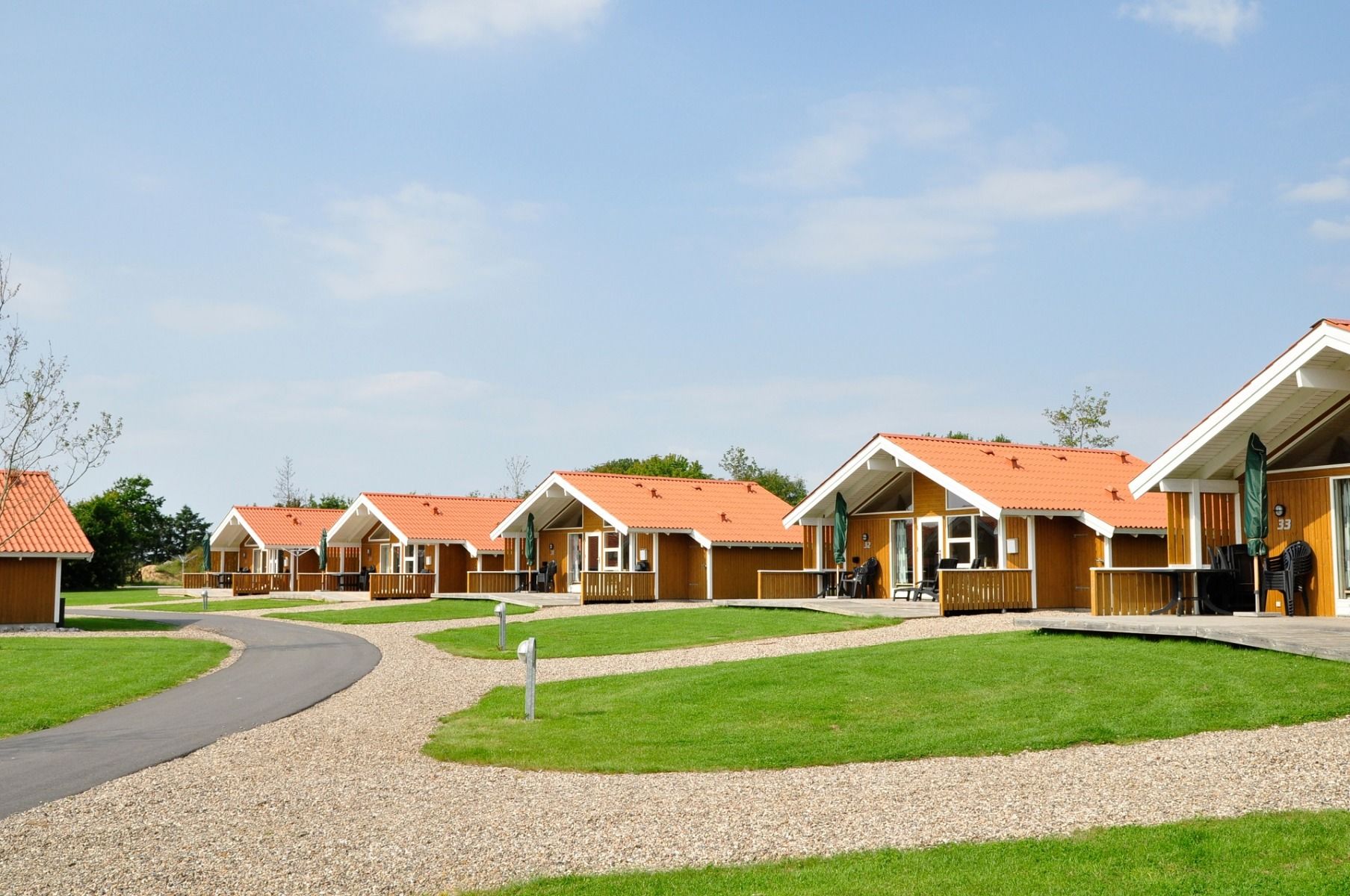 Eine Reihe kleiner, gelber Holzhütten mit roten Dächern säumt einen geschwungenen Weg im Sportzentrum Skaerbaek, umgeben von grünem Gras und Bäumen unter einem hellen, teilweise bewölkten Himmel.