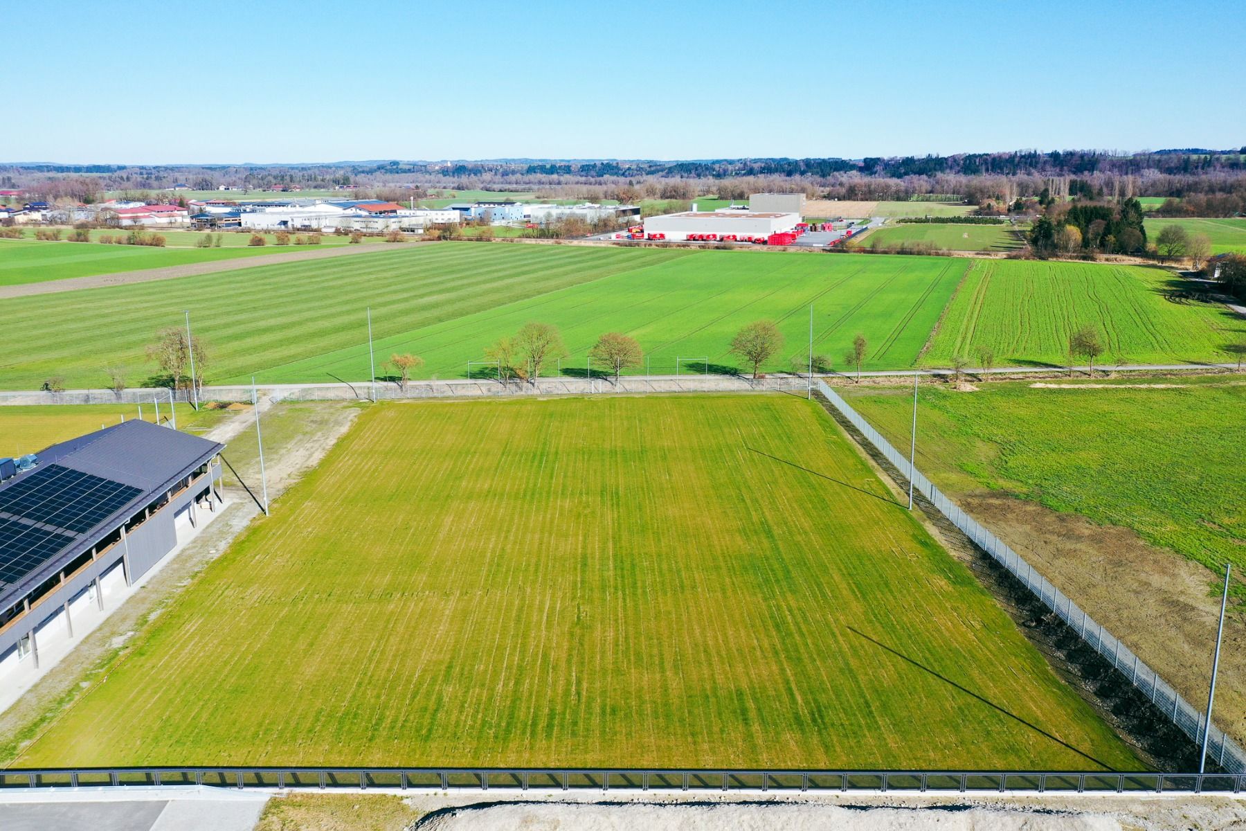 Blick auf grüne Wiesen, vereinzelte Bäume, Fußballplätze, ein Gebäude mit Sonnenkollektoren, weit entfernte Industriebauten und strahlend blauer Himmel - das Egger Stüberl ist das ideale Ziel für Ihr nächstes Trainingslager.