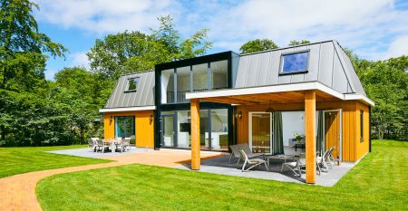 Modernes zweist&ouml;ckiges Haus mit Metalldach, gro&szlig;en Fenstern und gelben Holzverkleidungen. &Uuml;berdachte Terrasse mit Gartenm&ouml;beln und Essbereich mit Blick auf einen gr&uuml;nen Rasen - perfekt zum Entspannen nach einem Tag in Duinrell.