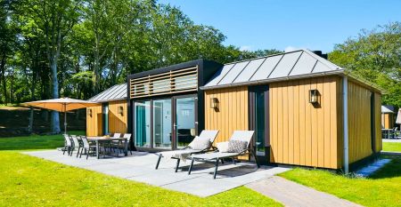 Modernes zweist&ouml;ckiges Haus mit Metalldach, gro&szlig;en Fenstern und gelben Holzverkleidungen. &Uuml;berdachte Terrasse mit Gartenm&ouml;beln und Essbereich mit Blick auf einen gr&uuml;nen Rasen - perfekt zum Entspannen nach einem Tag in Duinrell.