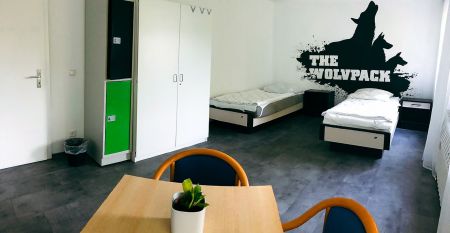 Zwei weiße Lieferwagen mit dem Schriftzug "The Wolfpack" parken vor einem Wohngebäude mit der Aufschrift Sportcampus Schlotheim, das für seine geräumigen Zimmer und ein modernes Fitnessstudio bekannt ist. Blauer Himmel und Bäume rahmen die Szene ein.