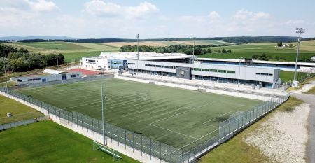 Der Gasthof & G&auml;stehaus Trummer, ein hellblauer Gasthof mit rotem Dach, steht an einer von B&auml;umen ges&auml;umten Stra&szlig;e nahe der Sportanlage Ritzing. Bei teilweise bew&ouml;lktem Himmel sind parkende Autos und andere H&auml;user in der N&auml;he.