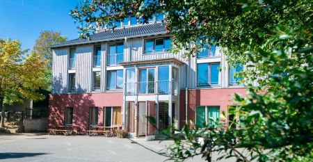 Das G&auml;stehaus Nettersheim ist ein modernes, dreist&ouml;ckiges Geb&auml;ude mit gro&szlig;en Fenstern, einer roten unteren Fassade, h&ouml;lzernen Obergeschossen und einem kleinen Balkon, eingebettet in eine gr&uuml;ne, nat&uuml;rliche Umgebung mit B&auml;umen unter der Sonne.