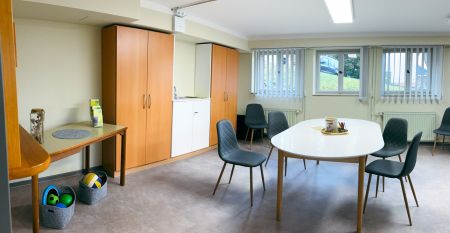 Das G&auml;stehaus Nettersheim ist ein modernes, dreist&ouml;ckiges Geb&auml;ude mit gro&szlig;en Fenstern, einer roten unteren Fassade, h&ouml;lzernen Obergeschossen und einem kleinen Balkon, eingebettet in eine gr&uuml;ne, nat&uuml;rliche Umgebung mit B&auml;umen unter der Sonne.