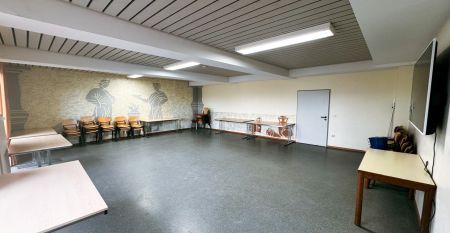 Das G&auml;stehaus Nettersheim ist ein modernes, dreist&ouml;ckiges Geb&auml;ude mit gro&szlig;en Fenstern, einer roten unteren Fassade, h&ouml;lzernen Obergeschossen und einem kleinen Balkon, eingebettet in eine gr&uuml;ne, nat&uuml;rliche Umgebung mit B&auml;umen unter der Sonne.