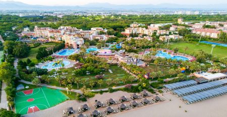 Luftfoto av Limak Arcadia Hotel med bassenger, hager, idrettsbaner og strandhytter - ideelt for fotballtreningsleiren i Tyrkia - omgitt av palmer og fjell under en klar himmel.