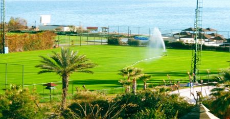 Luftfoto av Limak Arcadia Hotel med bassenger, hager, idrettsbaner og strandhytter - ideelt for fotballtreningsleiren i Tyrkia - omgitt av palmer og fjell under en klar himmel.