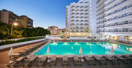 Hotelanlage bei D&auml;mmerung mit beleuchtetem Pool, Sonnenliegen und mehrst&ouml;ckigem wei&szlig;en Geb&auml;ude.