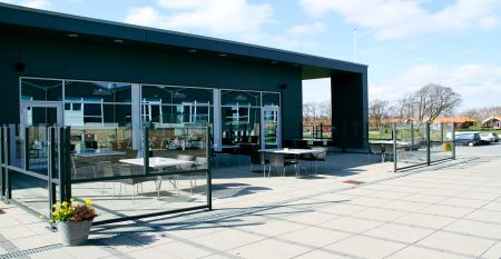 Eine Reihe kleiner, gelber Holzh&uuml;tten mit roten D&auml;chern s&auml;umt einen geschwungenen Weg im Sportzentrum Skaerbaek, umgeben von gr&uuml;nem Gras und B&auml;umen unter einem hellen, teilweise bew&ouml;lkten Himmel.