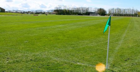 Eine Reihe kleiner, gelber Holzh&uuml;tten mit roten D&auml;chern s&auml;umt einen geschwungenen Weg im Sportzentrum Skaerbaek, umgeben von gr&uuml;nem Gras und B&auml;umen unter einem hellen, teilweise bew&ouml;lkten Himmel.